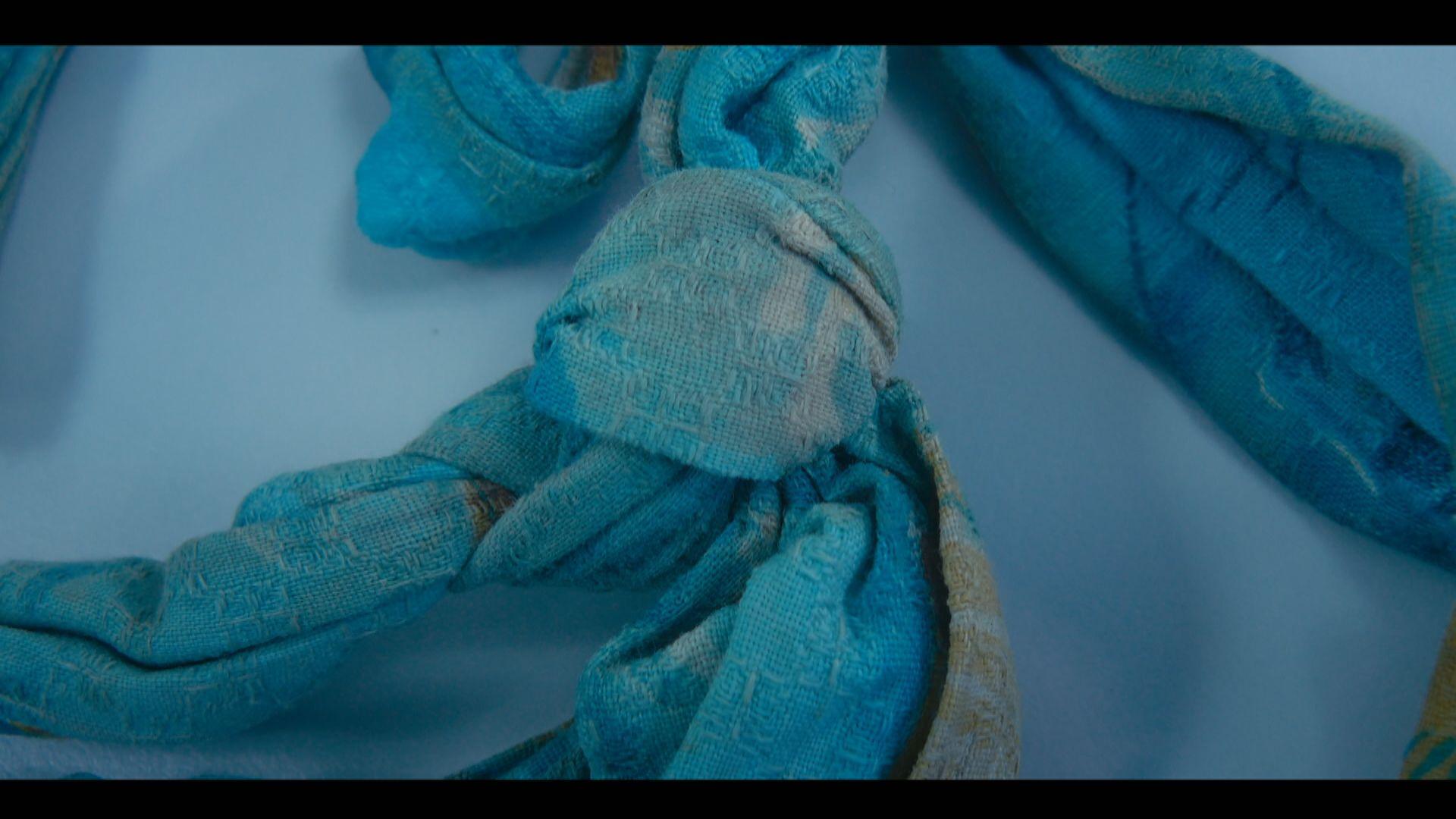 Close up of a knot in the grey/blue dressing gown cord used as a ligature to strangle Mary McLaughlin. The fabric is worn and slightly faded. It has been photographed against a light blue background.