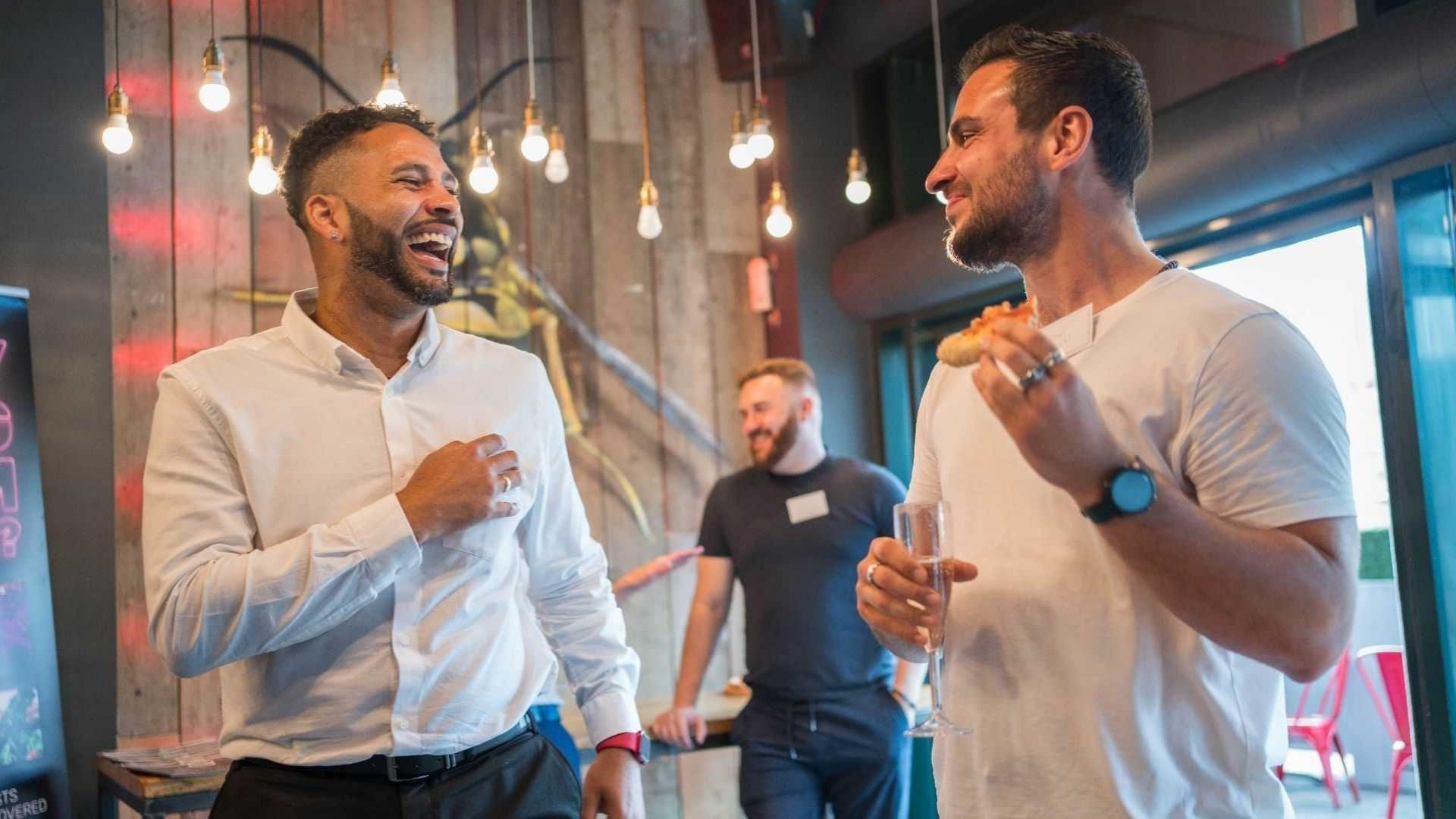Two men standing in animated conversation. The man on the left is laughing and dressed in a white buttoned-down shirt and black trousers. He is facing the man on the right who is smiling and is dressed in a white t-shirt and is holding a slice of pizza.