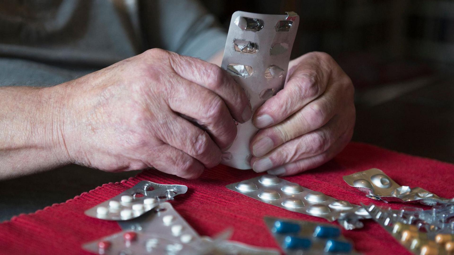 A close-up of hands holding a blister pack of tablets. There are several other packets of tablets laid out on a piece of red fabric in front of the person.