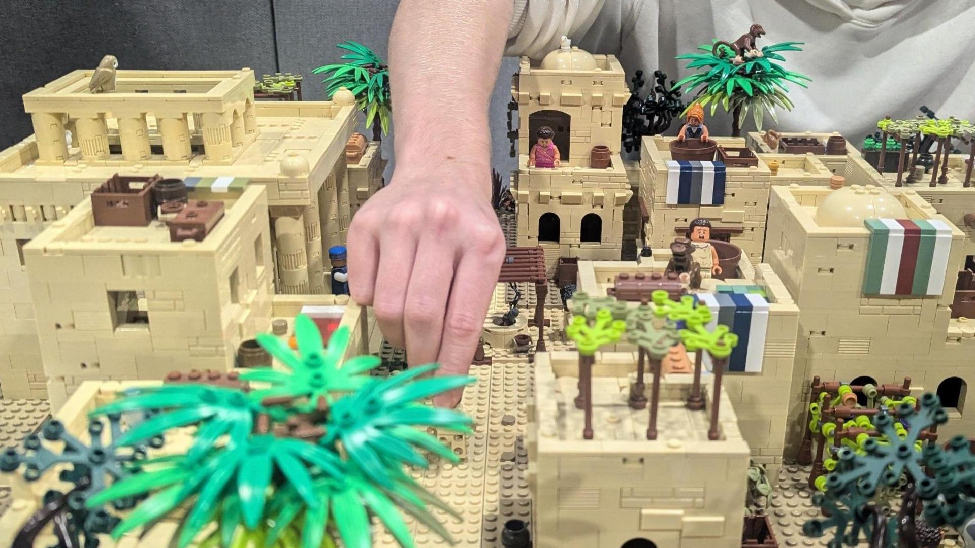 Mainly cream-coloured miniature model of ancient stone-built buildings with palm trees and a few people.  A hand is reaching down onto the model.