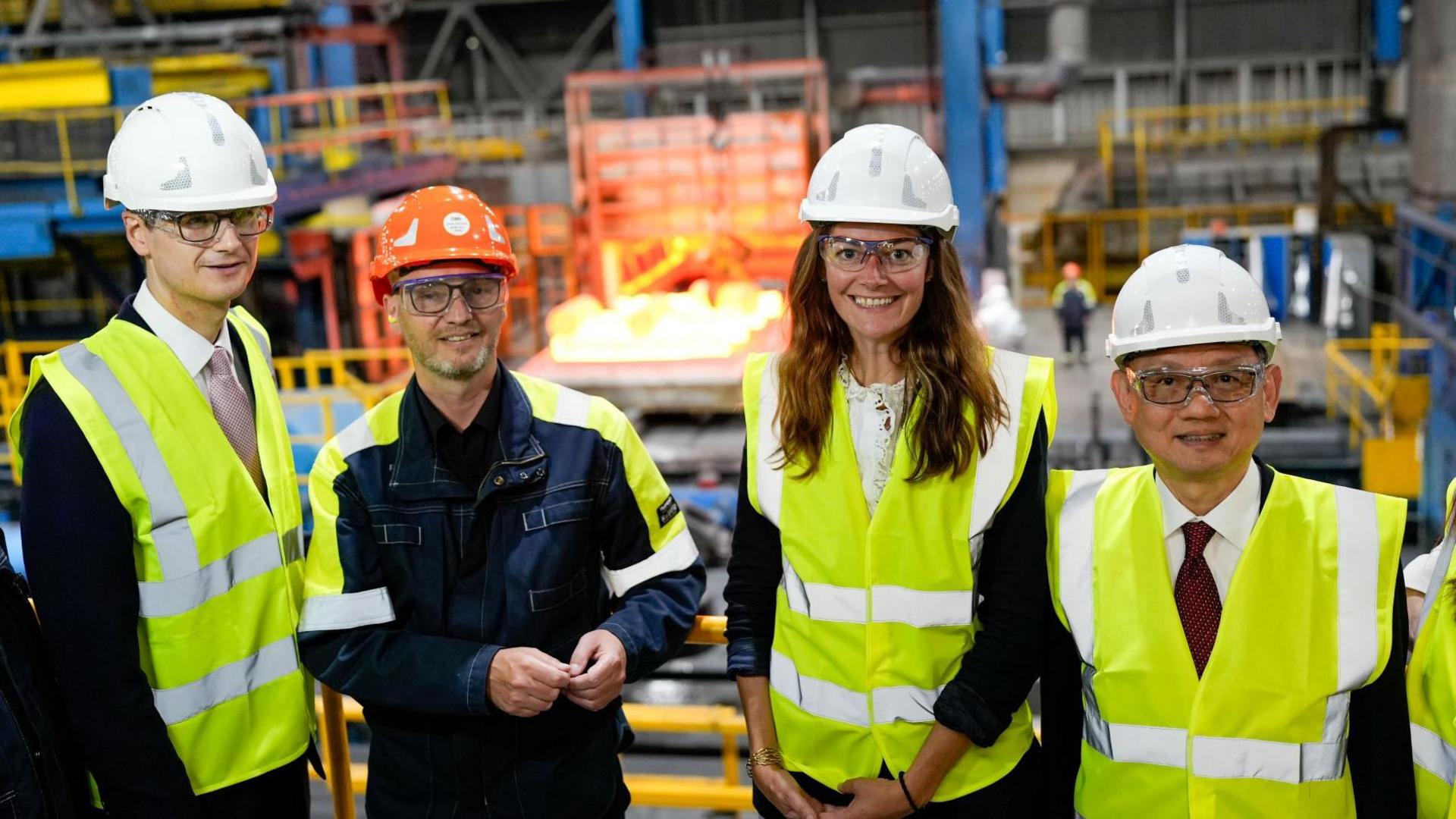 A group of three men and a woman stand in what appears to be a manufacturing plant. They all wear hi-vis jackets and helmets.
