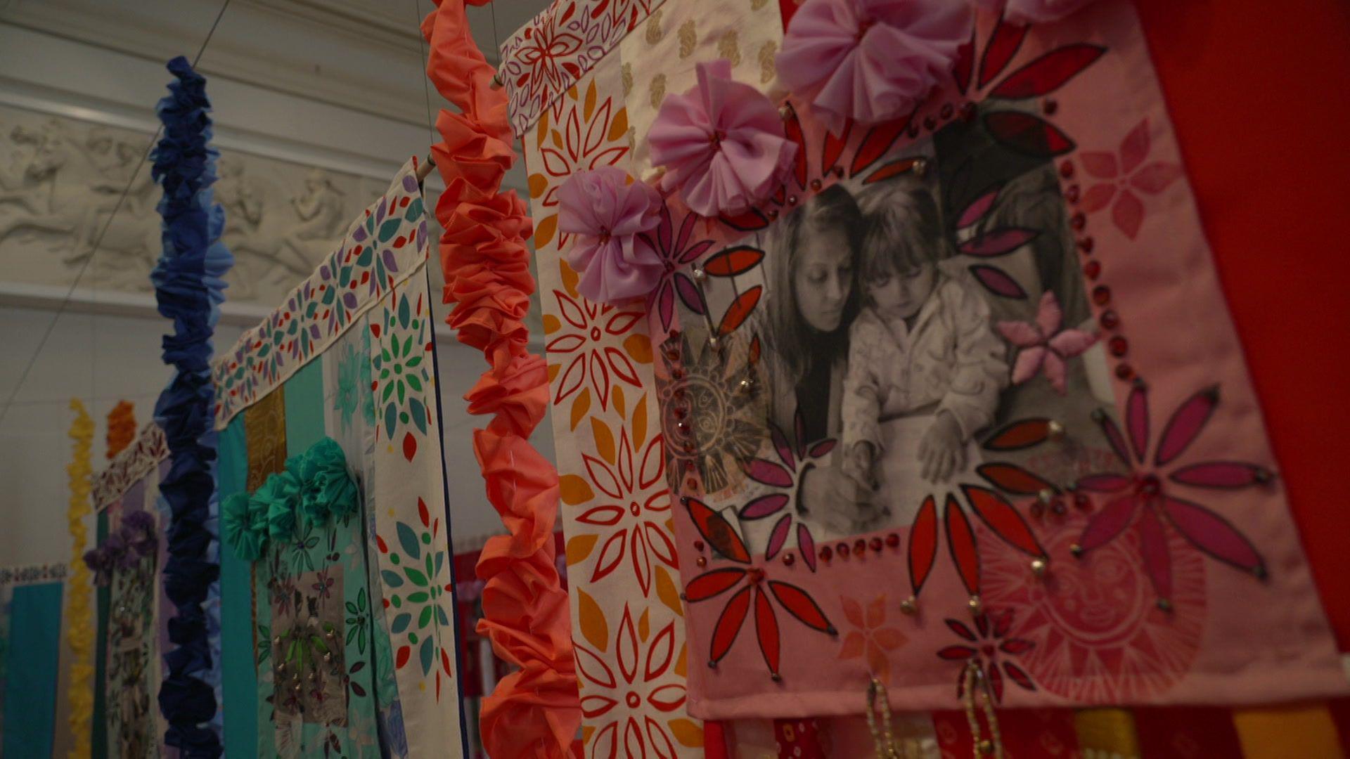 Pink, blue and yellow hangings showing a picture of a woman helping a girl to draw. They are adorned with floral petal designs and garlands.