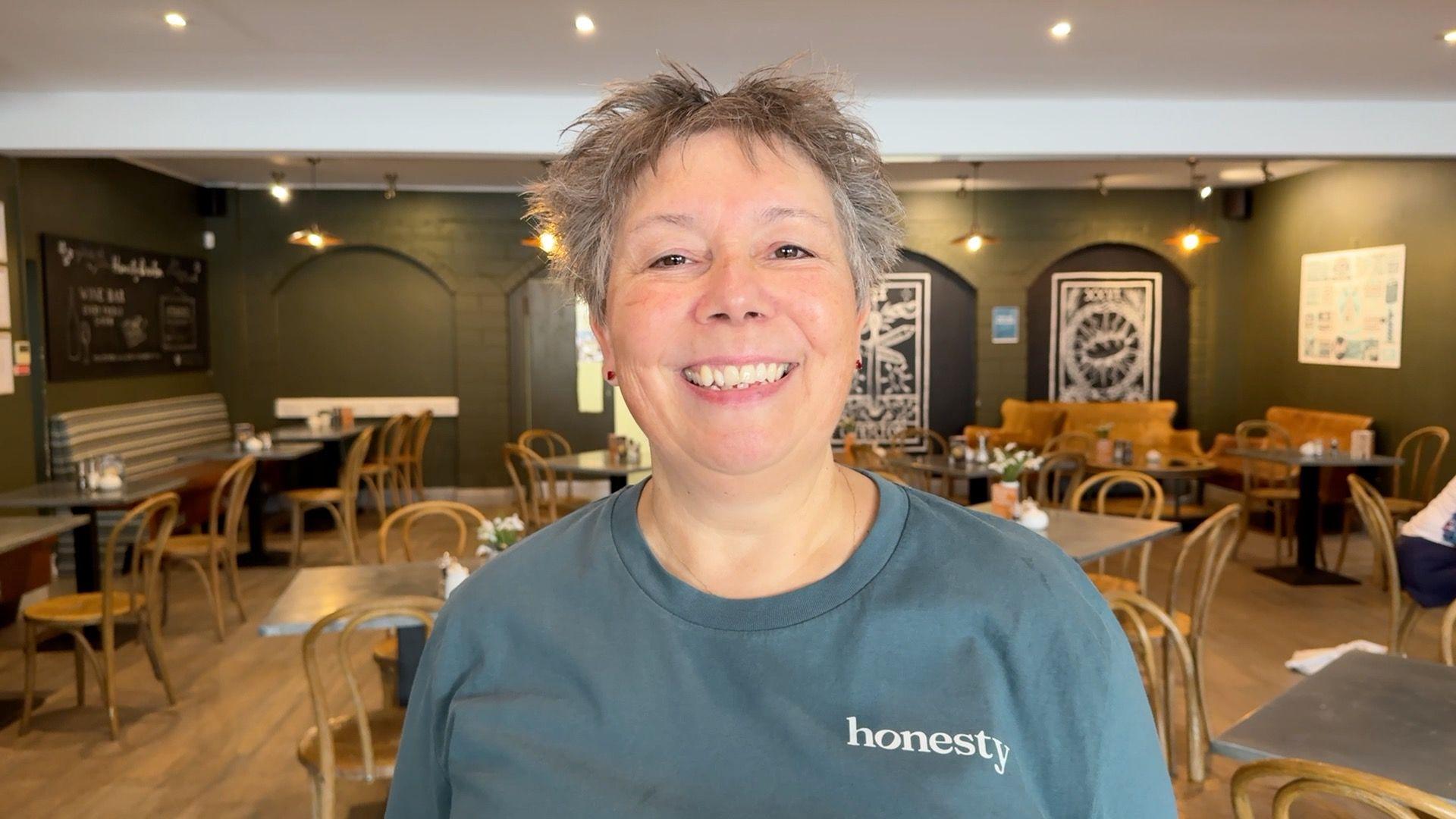 A woman with short grey hair and a bluey grey T-shirt, bearing the word "honesty" smiling and standing in a cafe