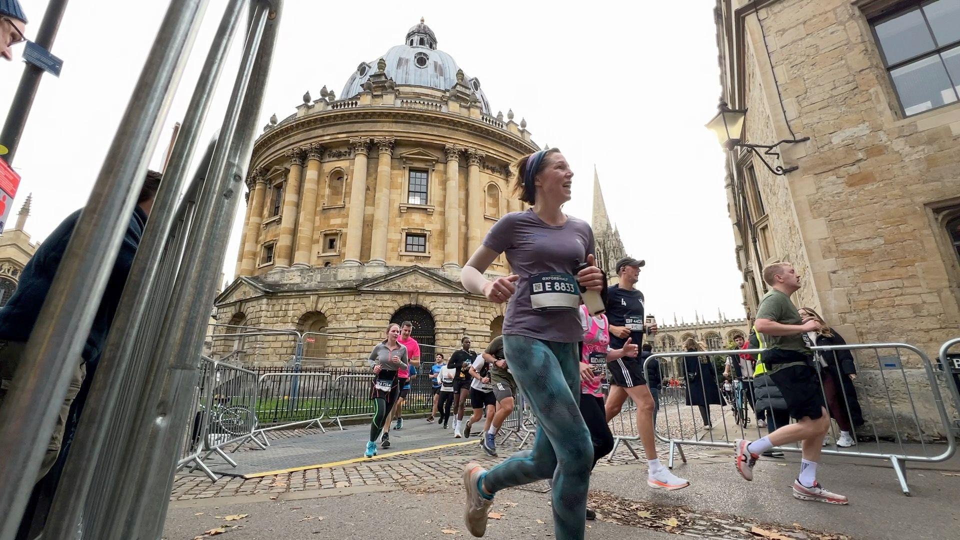 A group of people running between two gated supporter zones. Behind them is the Radcliffe camera.