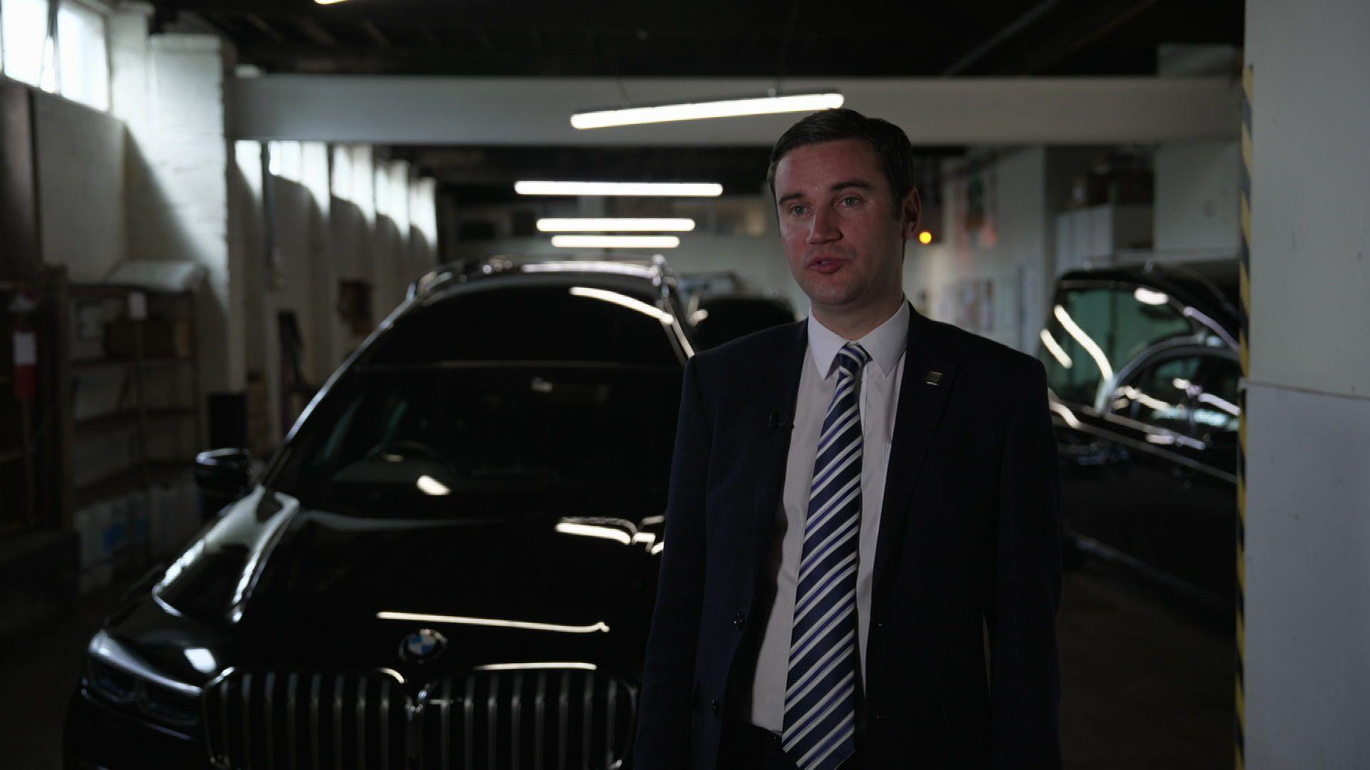 John Ware is standing in a large garage beside a black BMW hearse. He has dark hair and is wearing a dark suit, white shirt and striped tie.