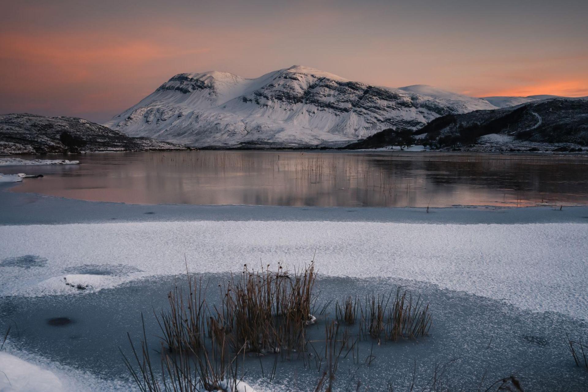 The mountain is covered in show and part of it is reflected in the water of a partly frozen loch. The sky has an orange hue to it.