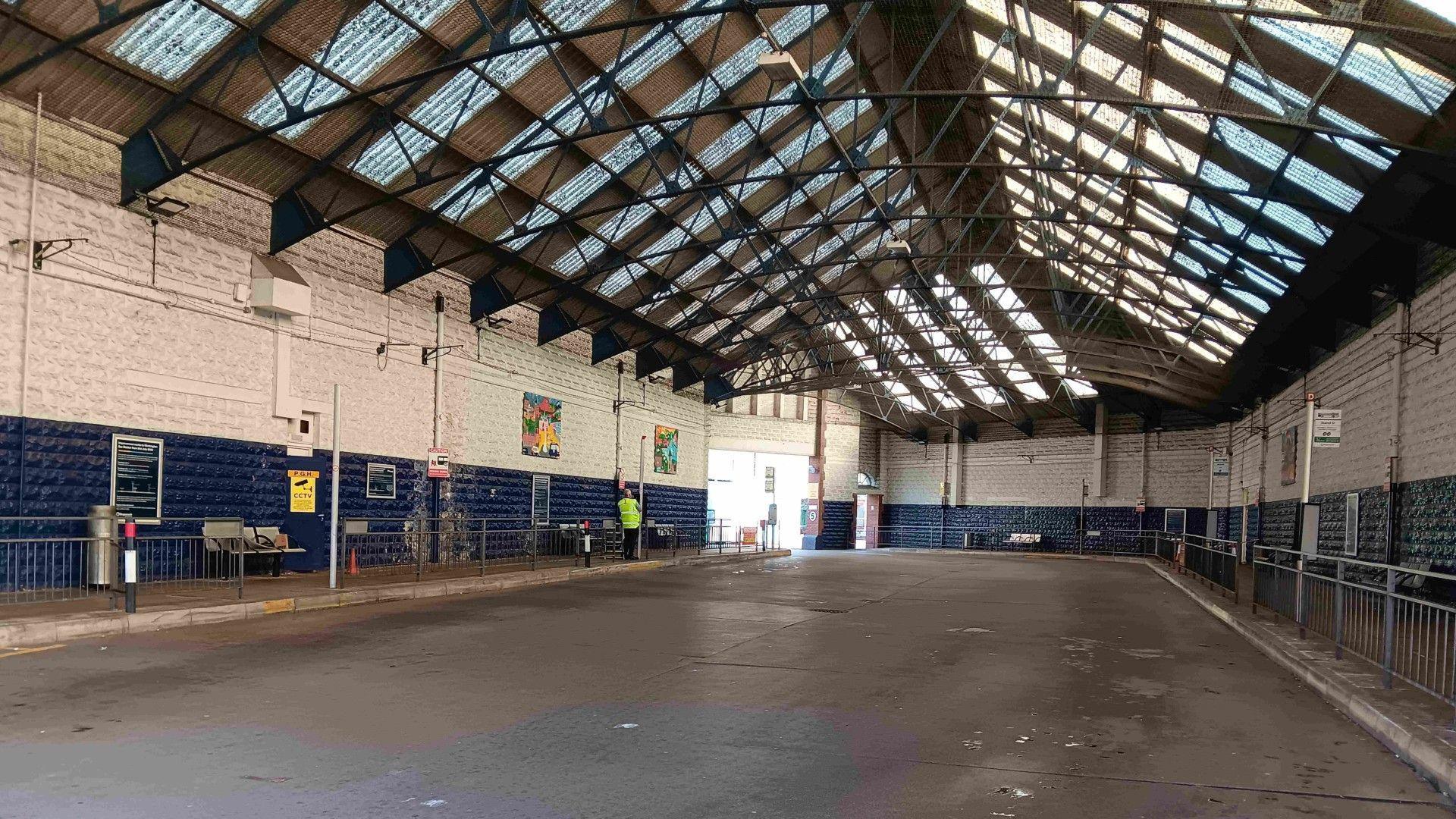 An internal shot of the old bus station. It has high ceilings with clear corrugated panels letting light in. There are bus stops at both sides of the concourse.