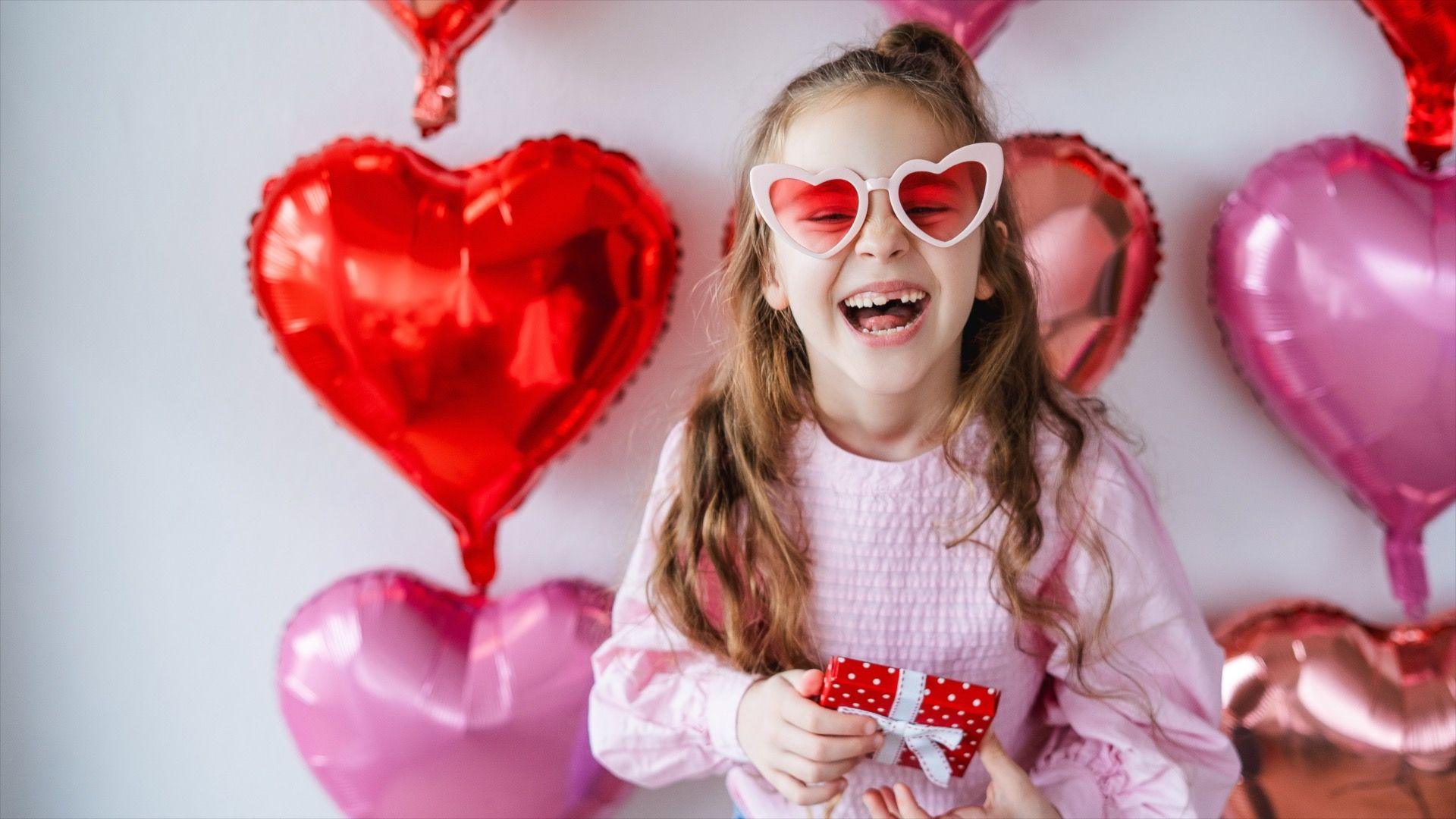 A girl wearing a pink top and pink heart shaped glasses is laughing as she stands in front of a white wall covered with red and pink heart shaped balloons. She's holding a present wrapped in red paper.