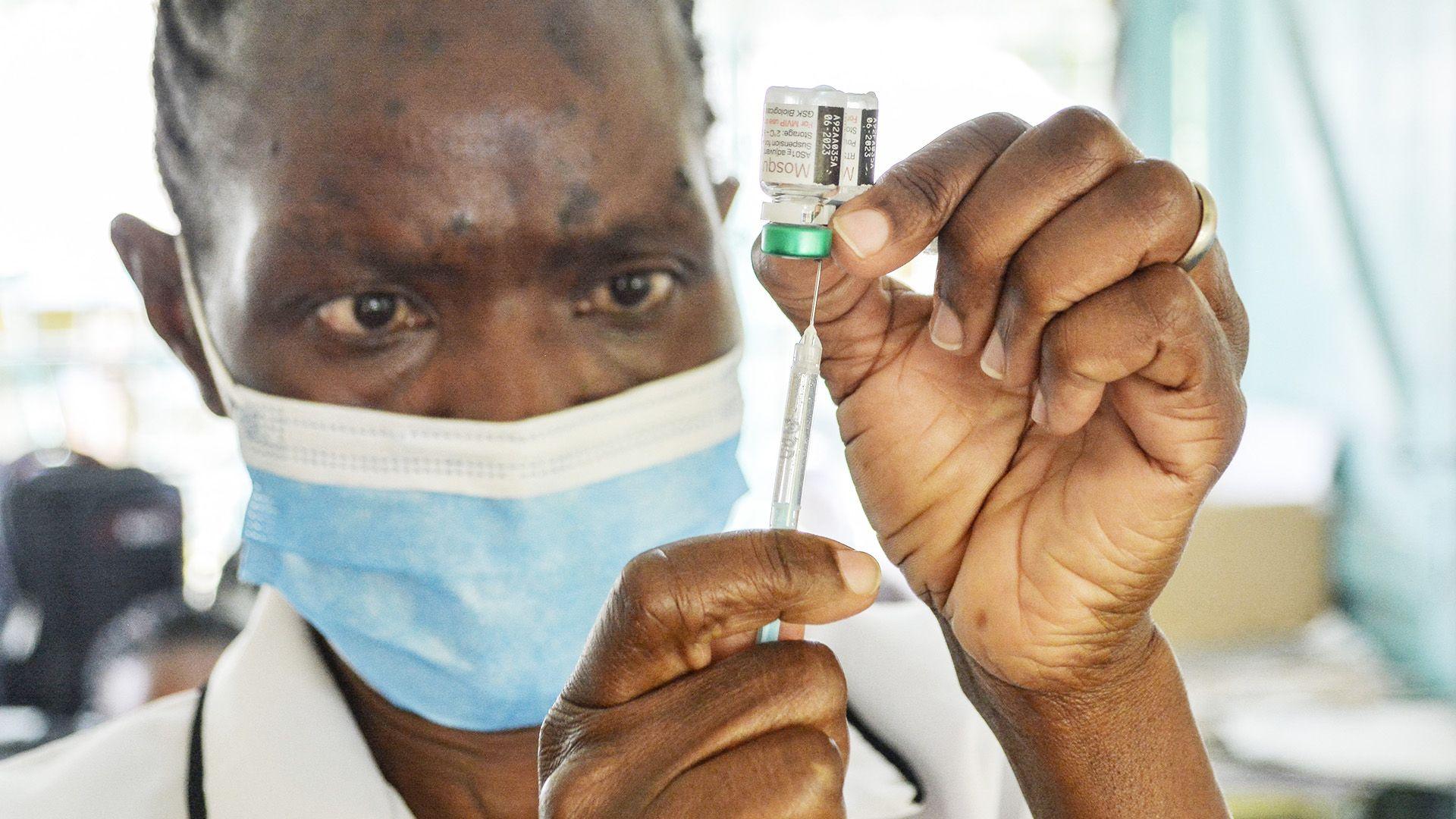 A person wearing a blue surgical mask prepares a malaria vaccine in a syringe.