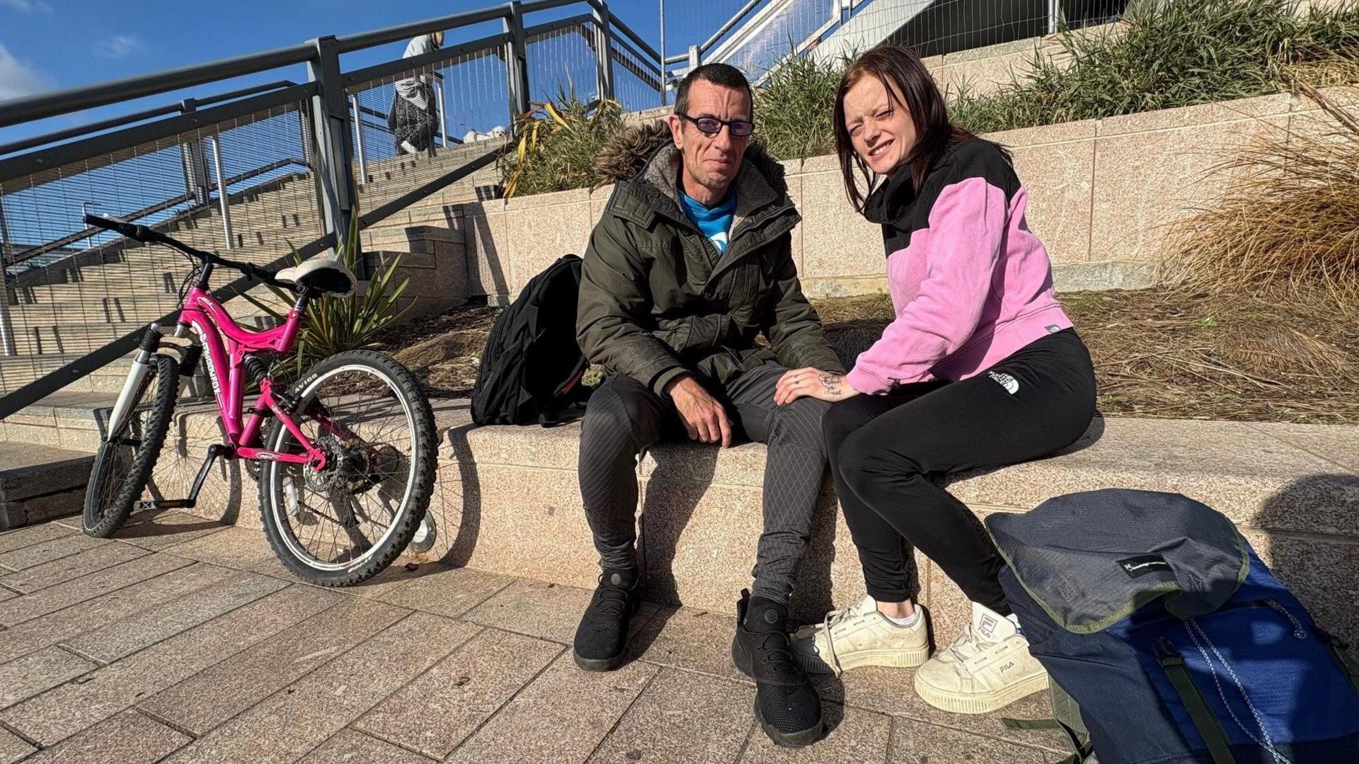 A couple, both with brown hair, are sitting on steps. Next to them is a bright pink bike.