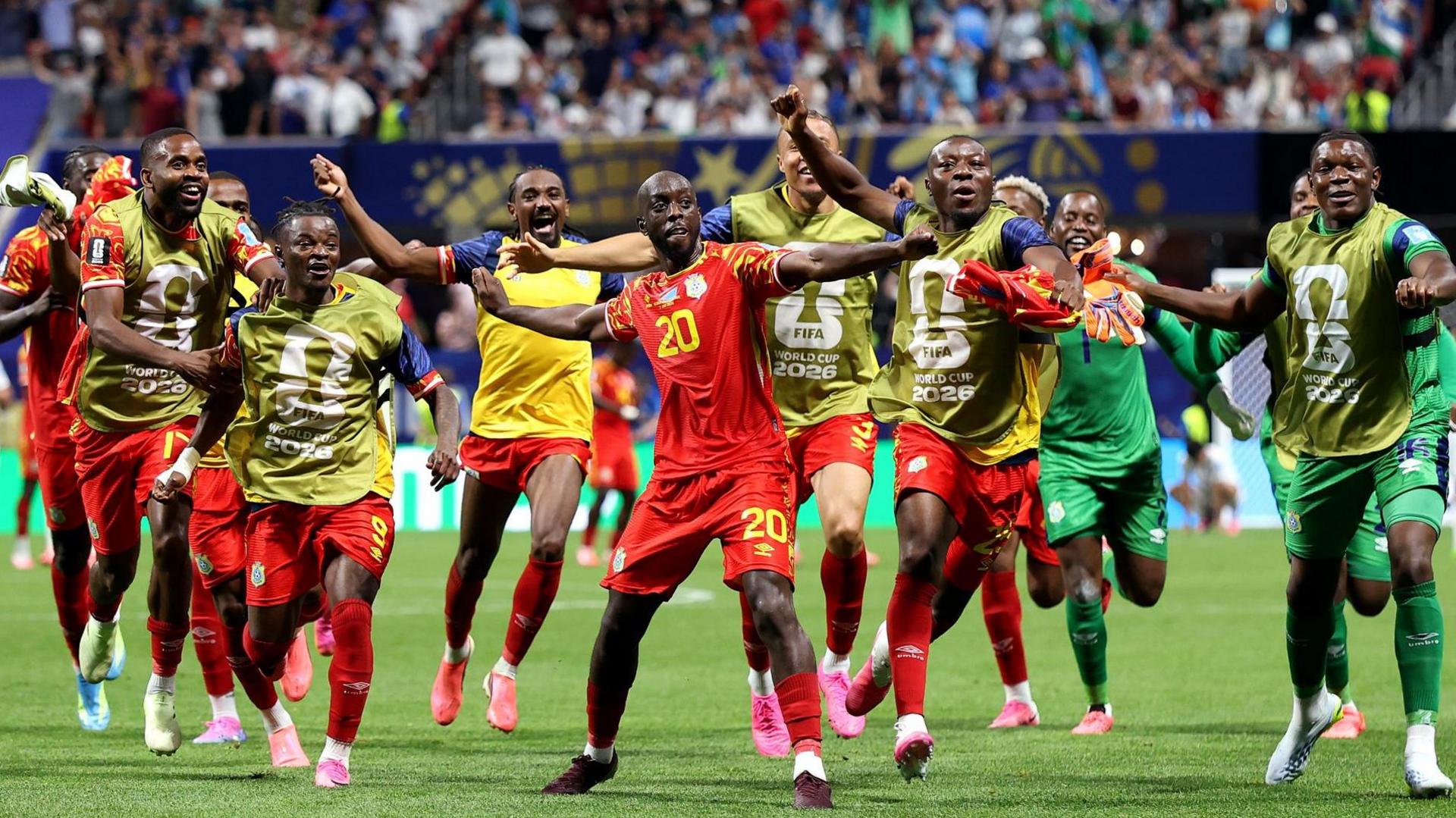 Yoane Wissa spreads his arms out as he dances in celebration after scoring a goal against Uzbekistan and the World Cup finals. He is surrounded by several DR Congo substitutes, who all bear smiles as some of them copy his pose