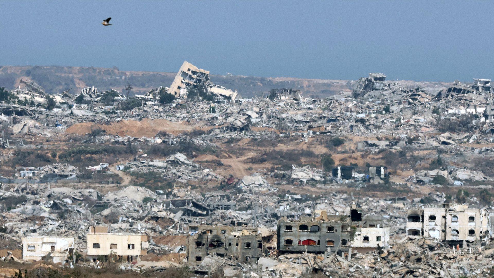 The image shows a vast area of destruction with numerous collapsed and damaged buildings. Rubble and debris are scattered across the landscape, indicating extensive devastation. In the foreground, there are partially standing structures with visible damage. A bird is flying in the sky above the ruins. The background features more destroyed buildings extending to the horizon under a clear blue sky.