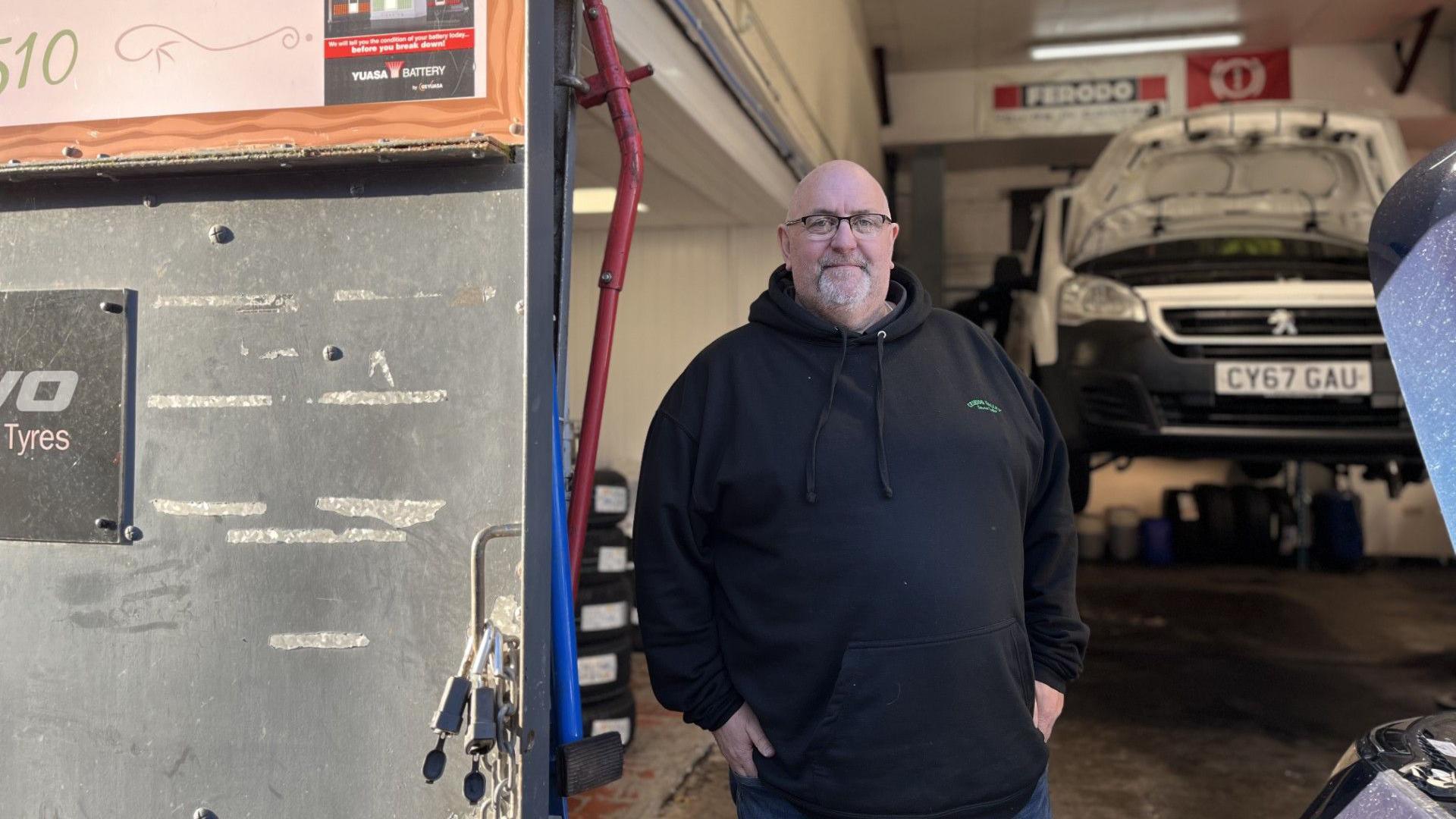 A bald man with a white goatee beard and wearing a black hoody and blue jeans. He is wearing black framed glasses and standing in a garage workshop.  There is a white car raised up behind him with the bonnet up.