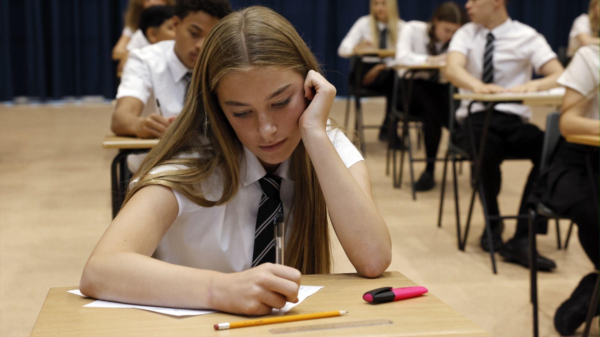 A girl wearing a white shirt and a dark blue tie sits at a desk in an exam hall. 