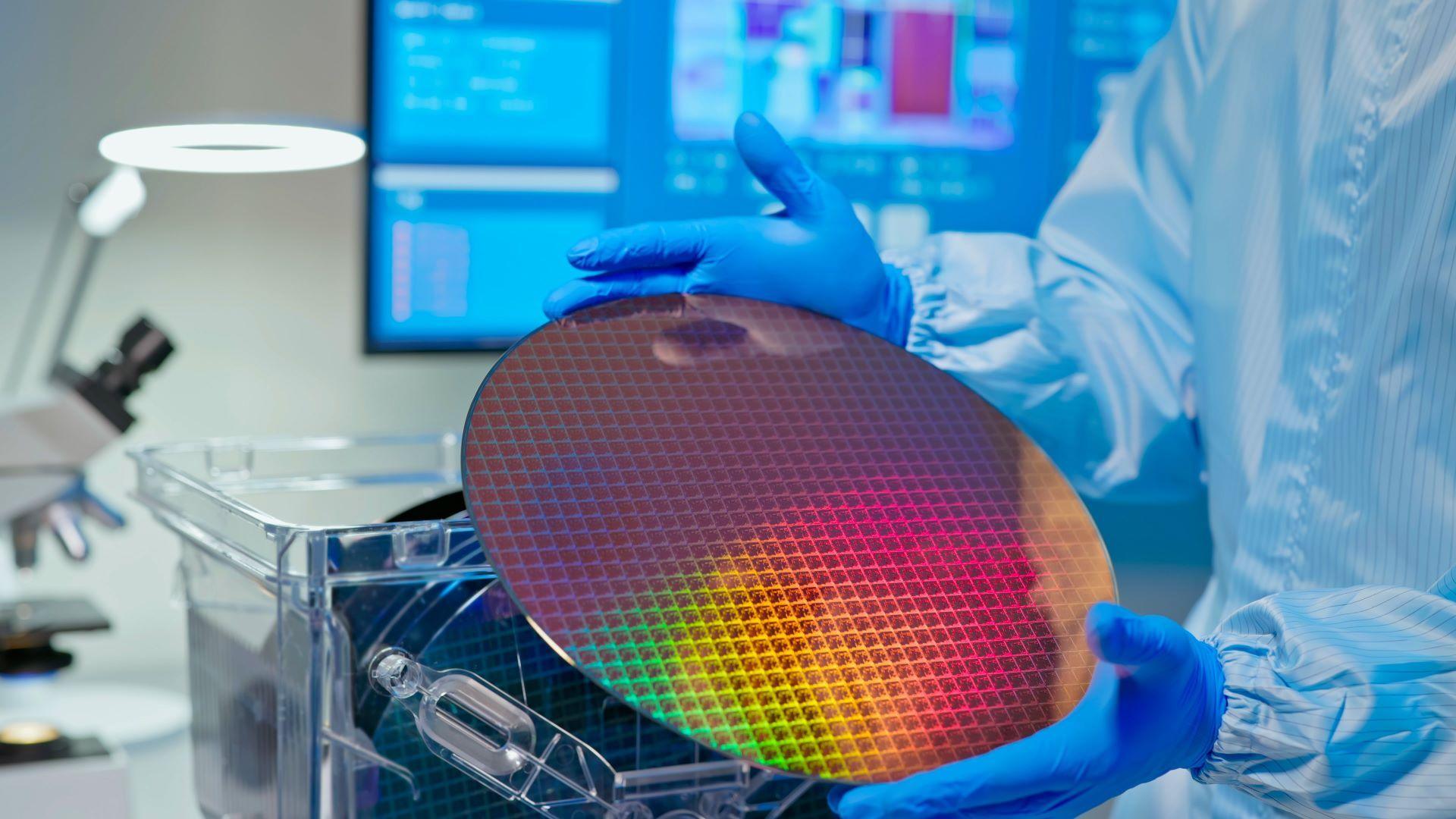 A technician holds up a silicon wafer - a round flat disc reflecting pink, red, yellow and green colours. 