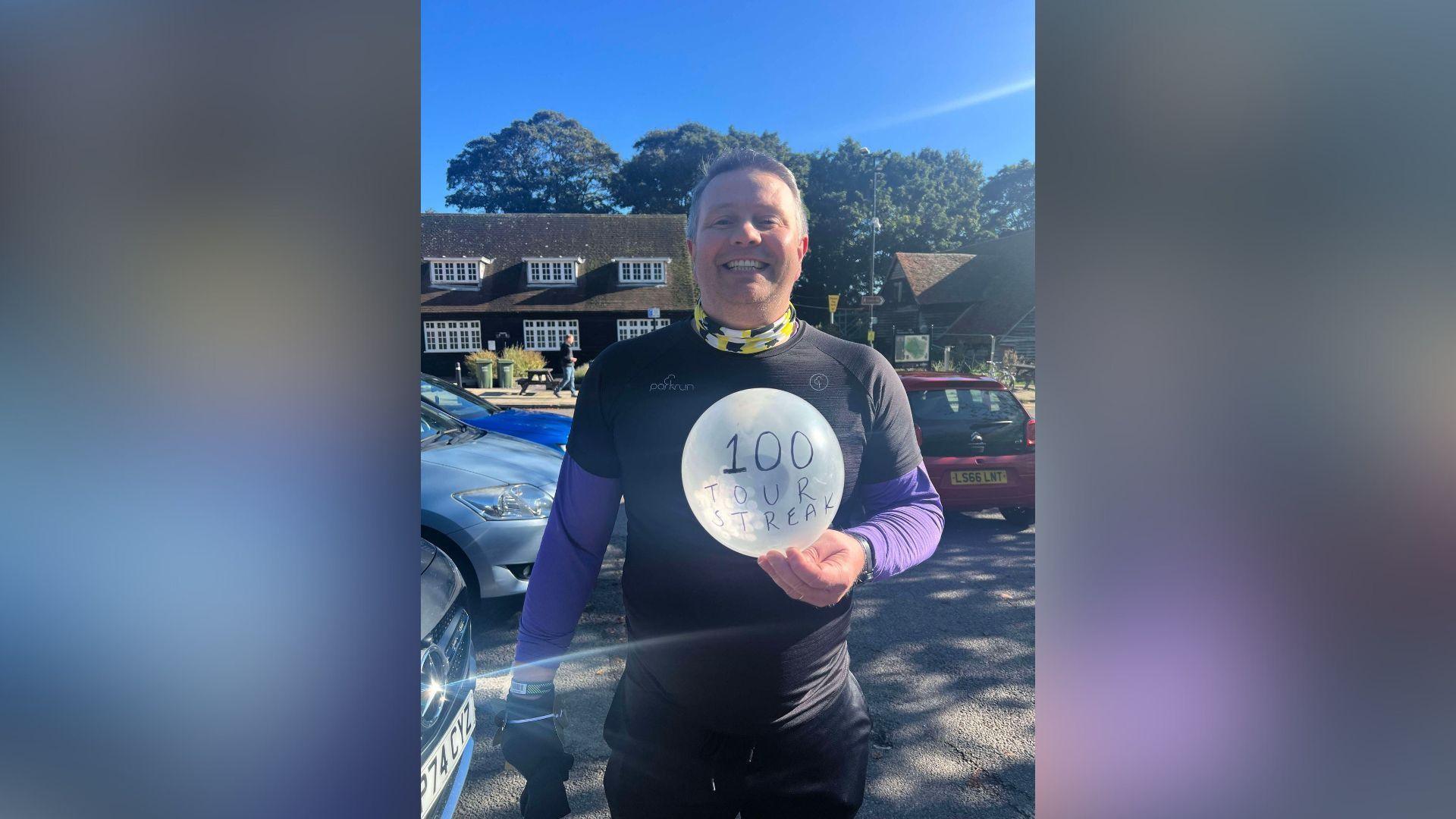 A smiling Ian Parris looking straight at the camera holding a balloon with the words: "100 tour streak". He is wearing a black and purple top and there are cars and a house in the background.