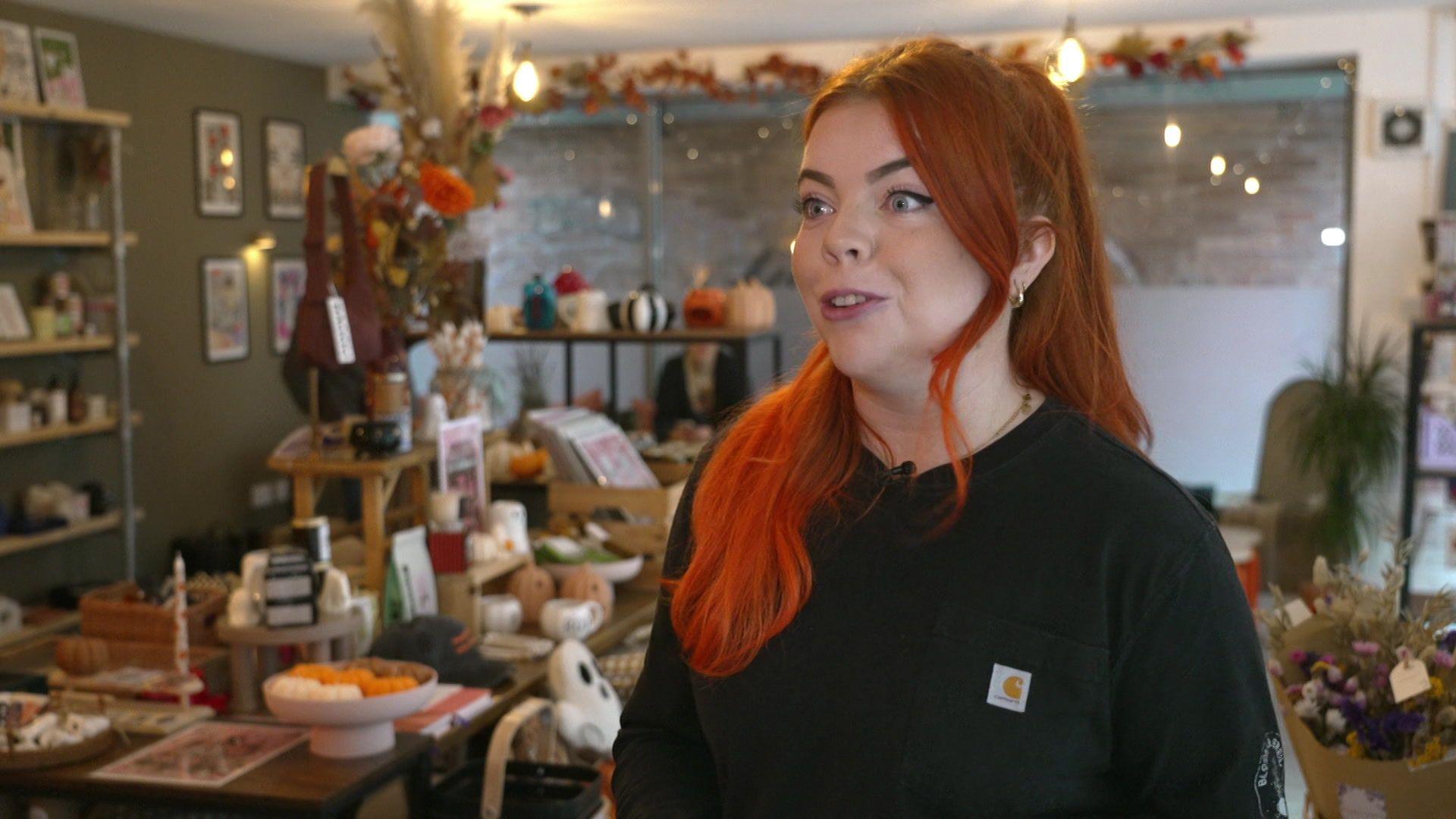 Charlotte Brennan with long, bright orange hair and a black sweater stands in front of her gift shop items which includes homeware,ghost-shaped pillows, dried flowers, candles and ornaments