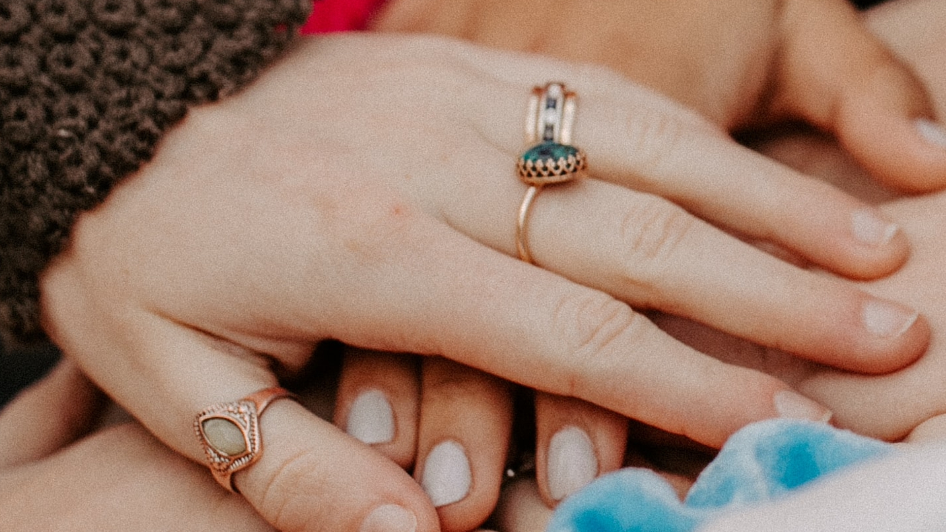 A close-up of two hands holding each other in a supportive way. The hand on top is wearing three rings and the hand below is wearing white nail varnish.