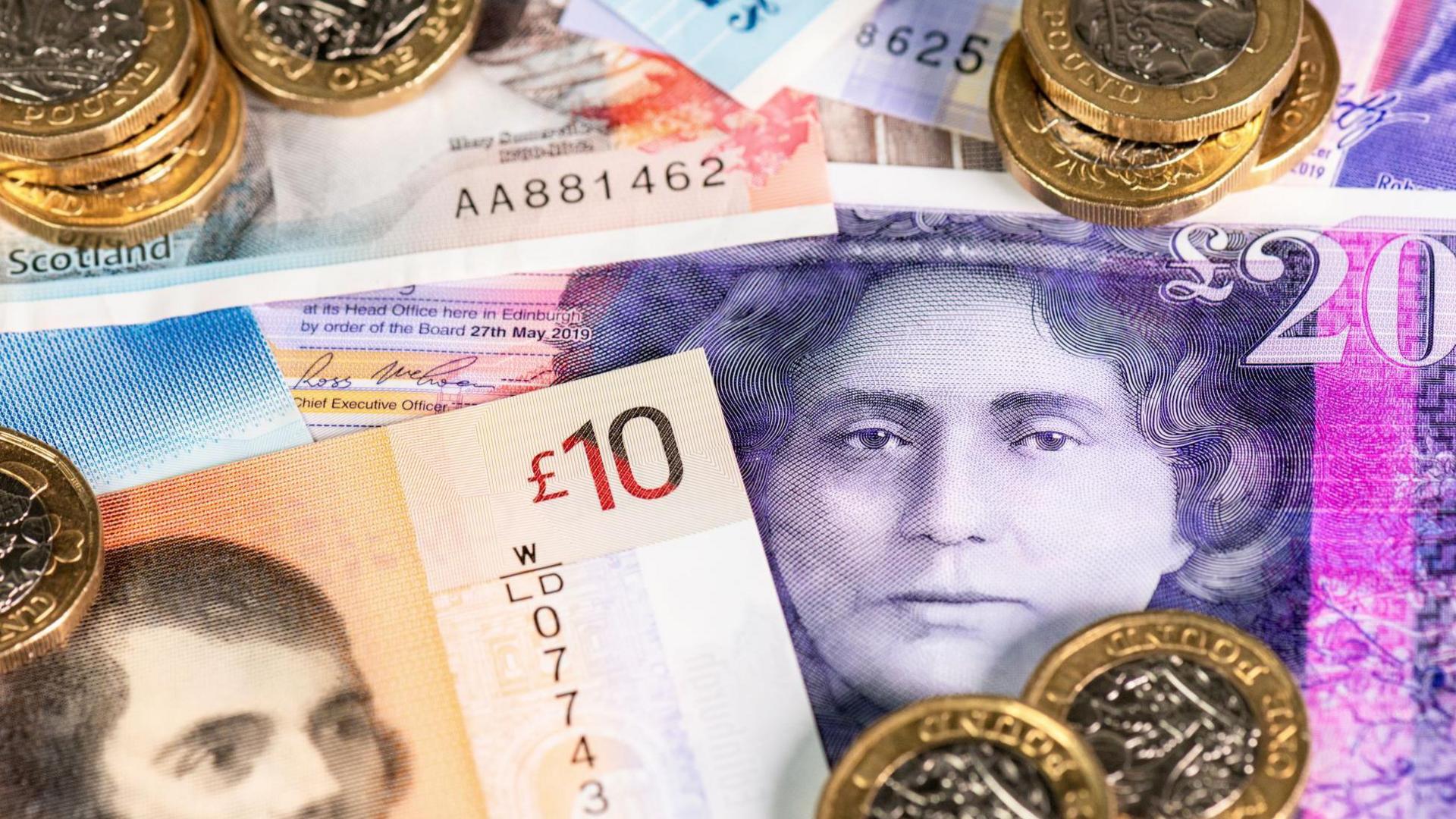 A collection of banknotes of different denominations, including £10 and £20. The colours of the notes include brown, pink and purple. On top of the banknotes are £1 coins.