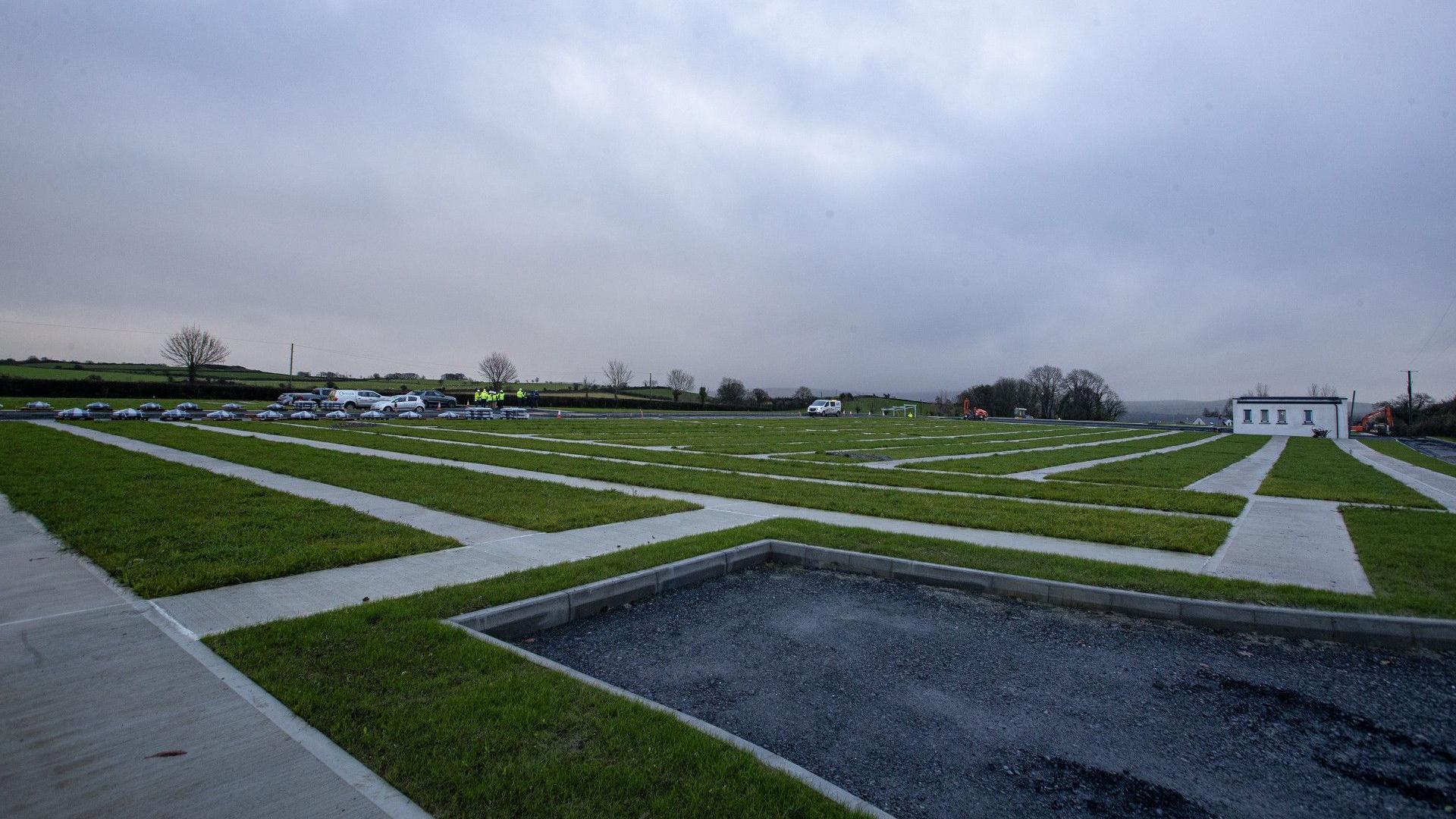 In the foreground there is a rectangle of tarmac, beyond which stretches a grassy expanse that is divided by lines of concrete paving. Far in the distance vans and other vehicles can barely be made out.