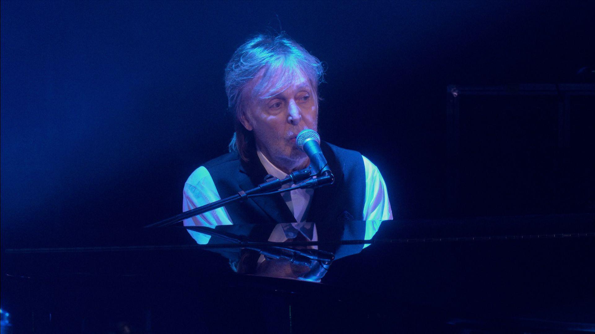 Sir Paul McCartney sings as he plays a grand piano, bathed in blue light