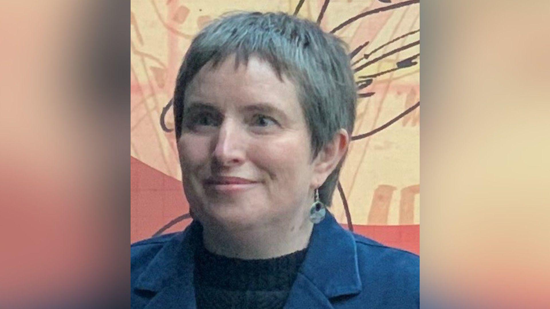 A woman with short grey hair, earrings, and wearing a blue jacket and dark top, smiles slightly as she looks to our left.