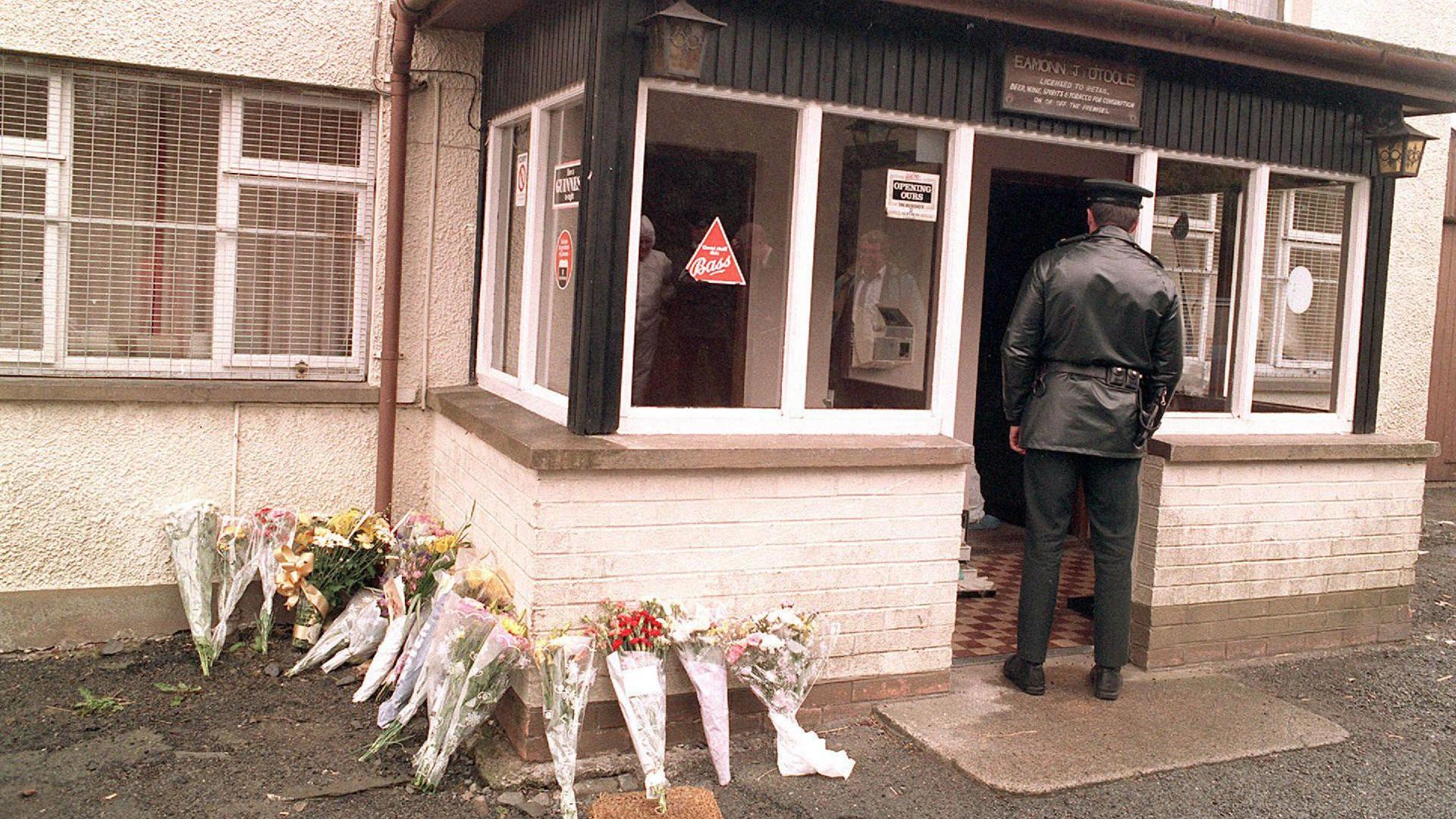 Archive image of Loughinisland massacre. A policeman is standing in the entrance of the building with a bunch of bouquets of flowers placed to the side.