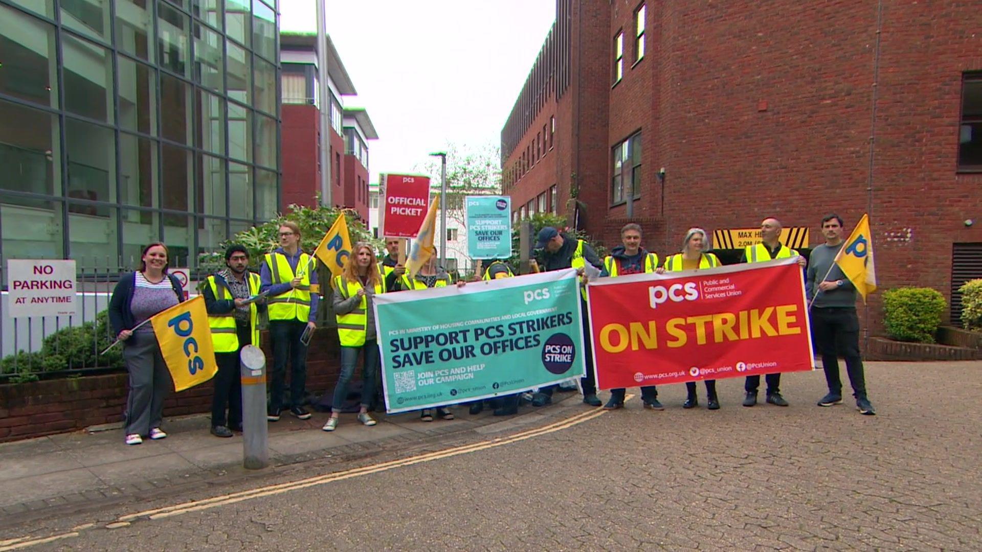 Workers stand on a road as part of a strike. The staff are wearing hi-vis jackets. They are holding flags and banners. One sign says "support PCS strikers save our offices".