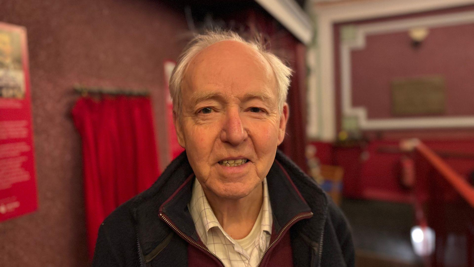 A man smiling straight at the camera. He is wearing a burgundy jumper, white shirt, and black jacket.