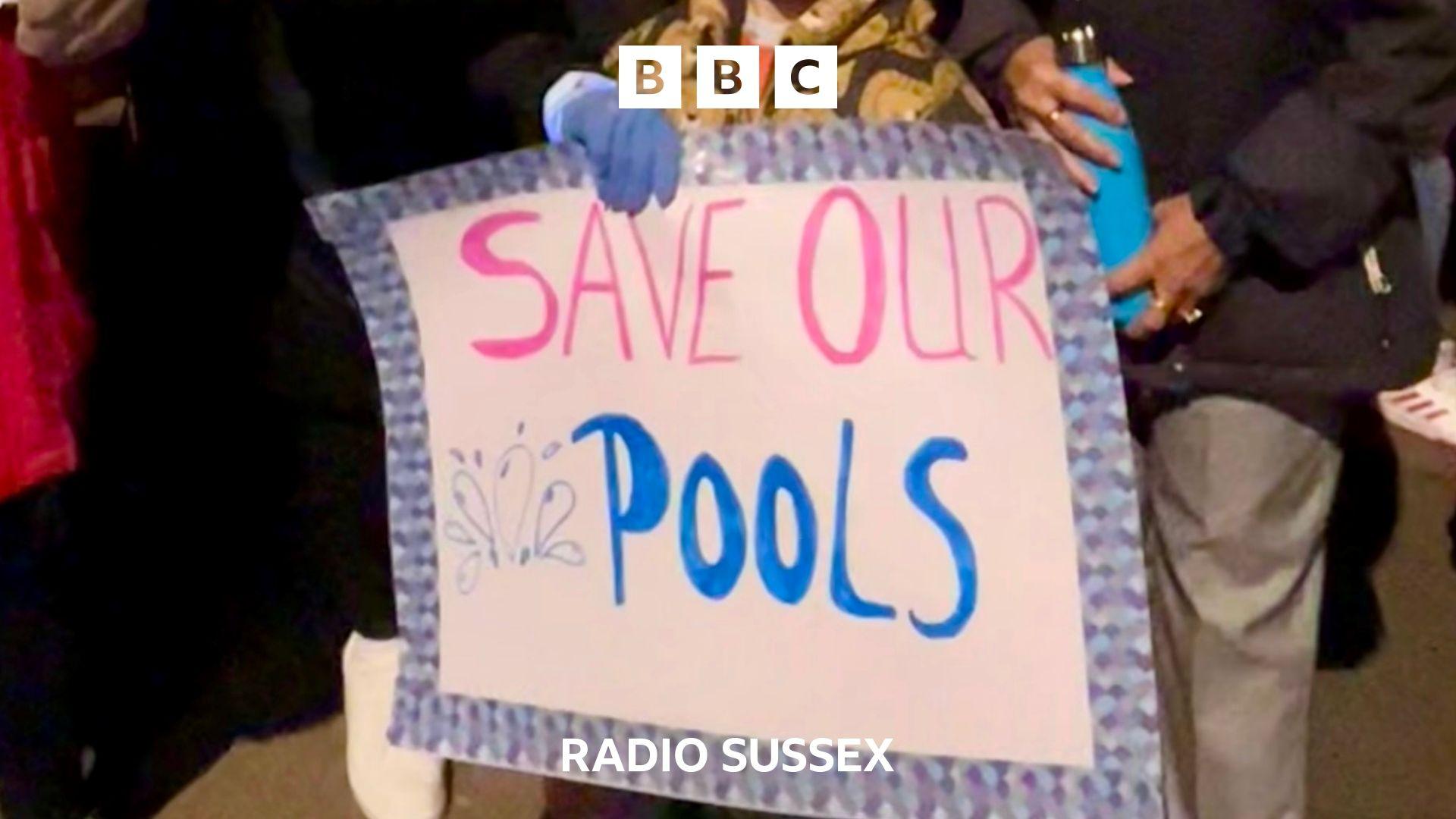 A gloved hand holds a hand drawn banner that says "SAVE OUR POOLS", in pink and blue text.