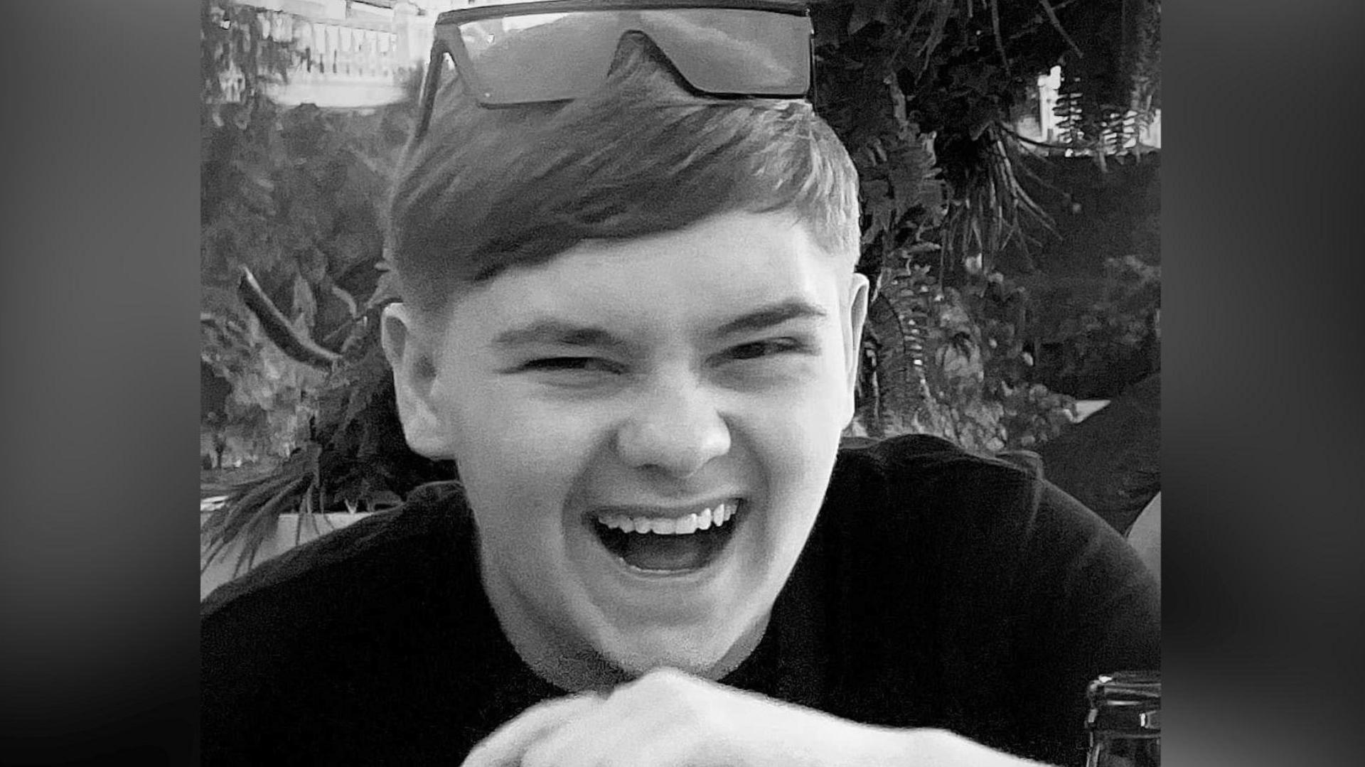 A black and white picture of the teenager who is laughing. He wears sunglasses on the top of his head, trees and plants can be seen in the background.