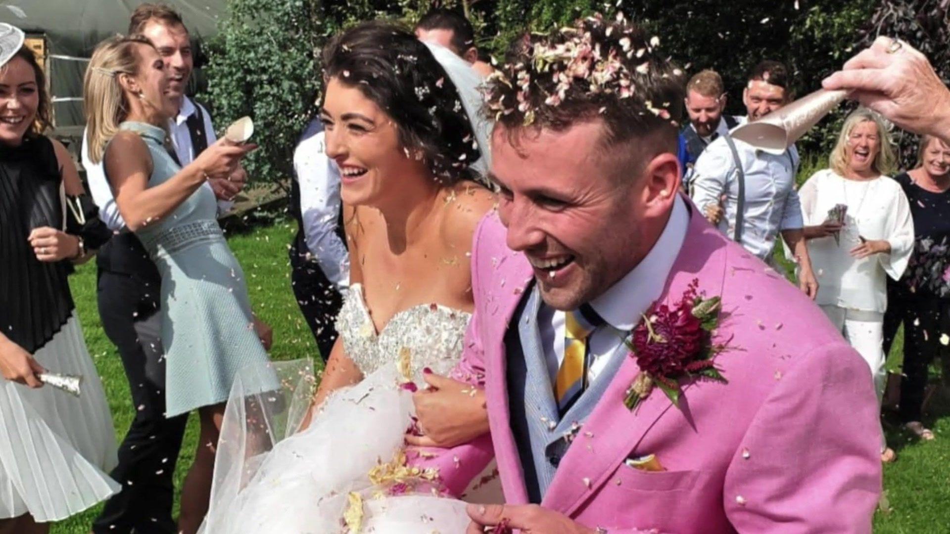 Sam and Holly Morgan on their wedding day. He is wearing a pink jacket and she has a strapless dress. They are both laughing and there is confetti in their hair.