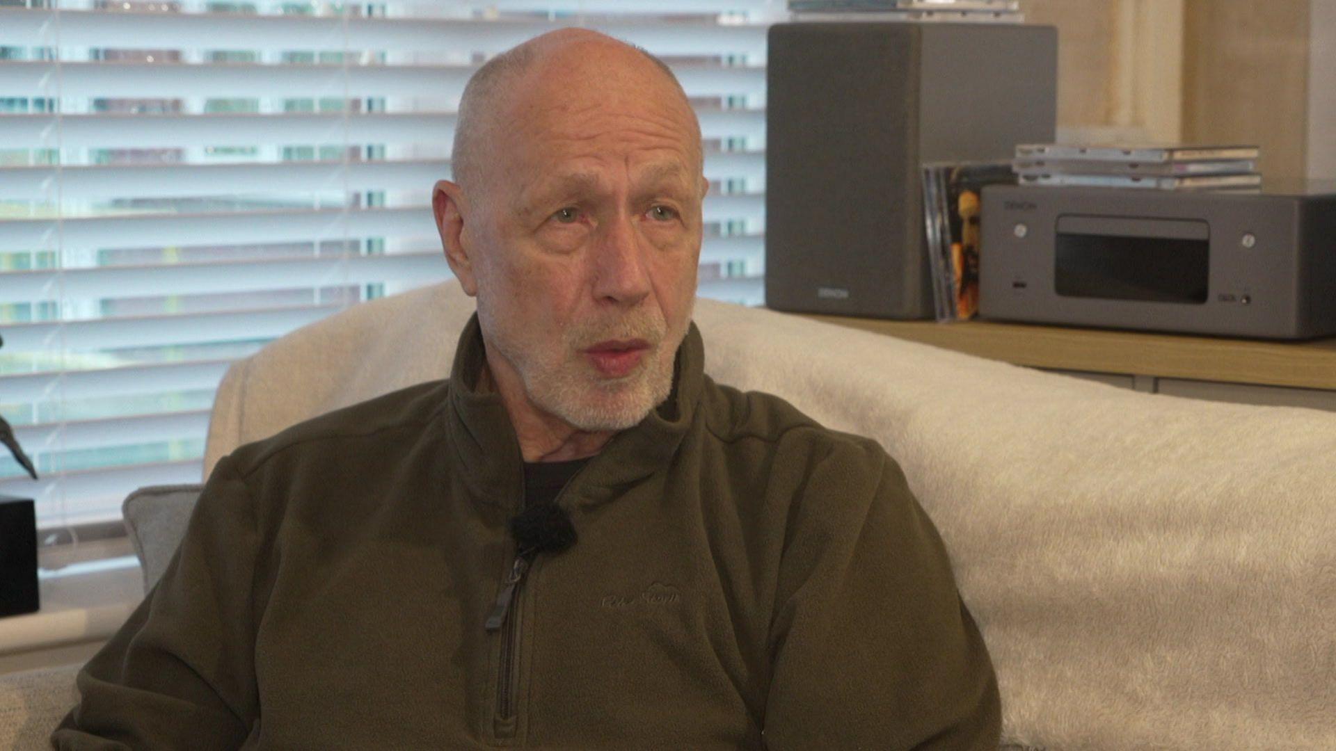 Dave Hemmings, who is bald and has a short grey beard, is sitting on the sofa in his bungalow. 