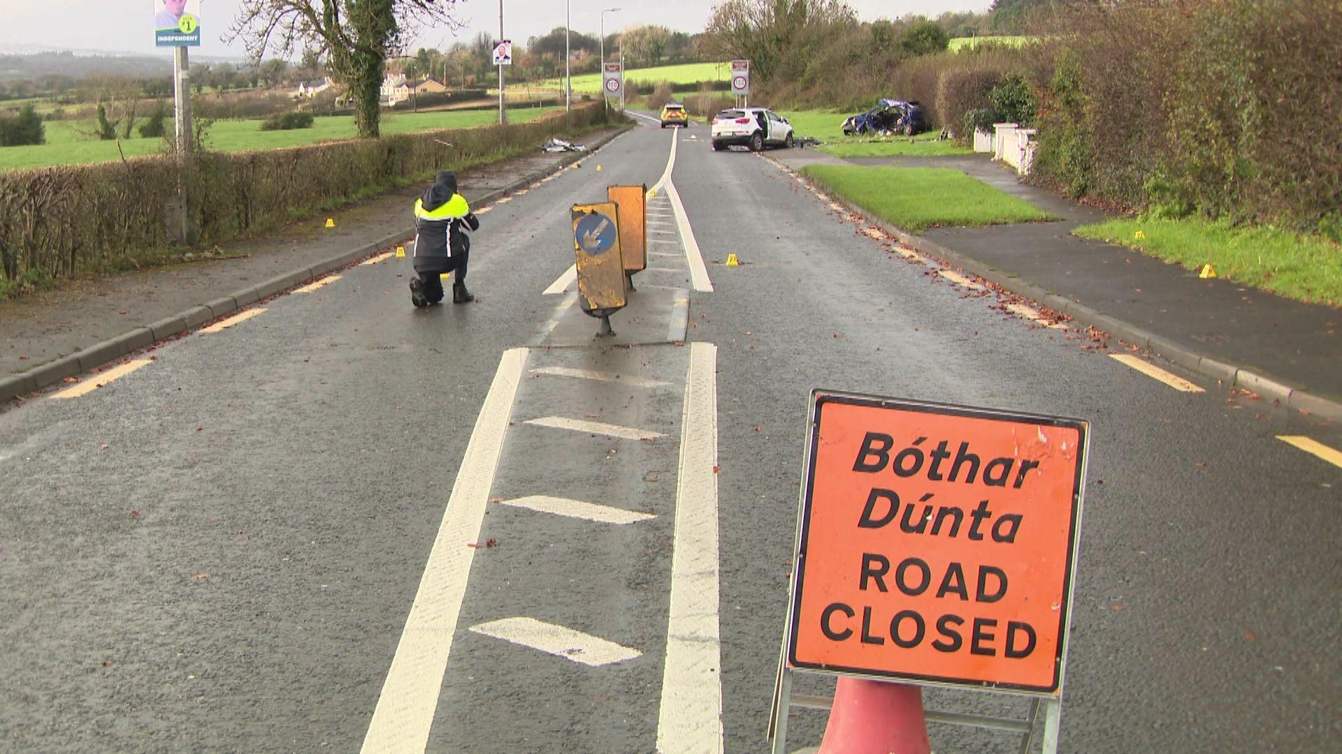 Two cars are visible in the rear of the picture. The remains of a blue car is on the grass.  A second white vehicle is on the road. A road closed sign is in the foreground