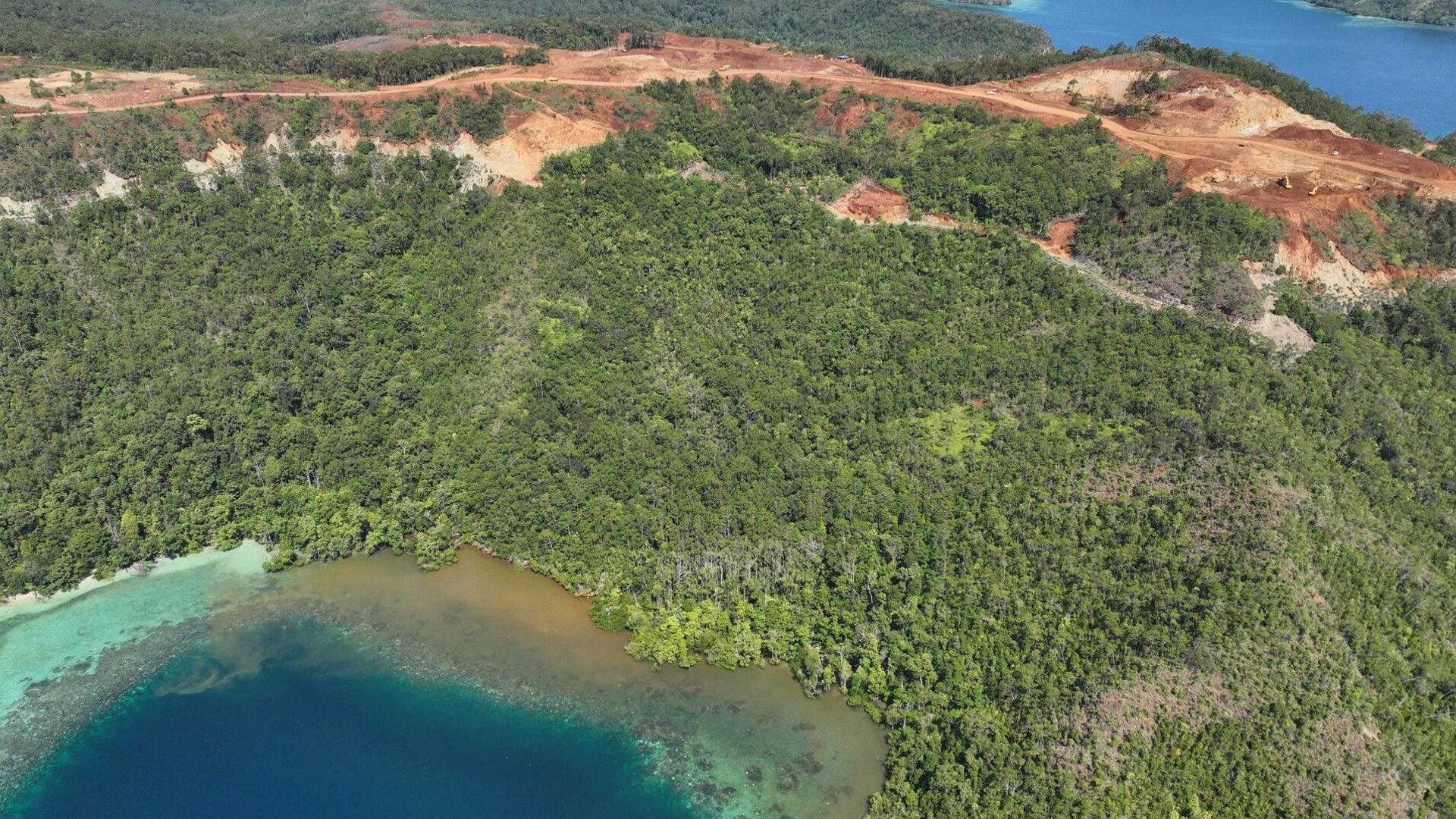 A photograph of mining on Kawei island in Raja Ampat, appears to show sediment  running into the coastal water. The aerial image shows a green, verdant island from above. Mining operations just uphill of the water's edge are in contrast to the lush forest - land has been cleared and brown earth is exposed. Downhill of the mine, brown-coloured sediment appears to be running into the clear, blue water. 