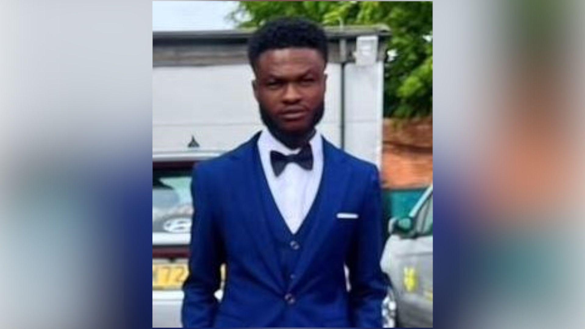 Clinton Antwi with medium-length dark hair and slight beard, wearing a blue jacket and black bow tie. He is standing outside next to a car and in front of a white building.