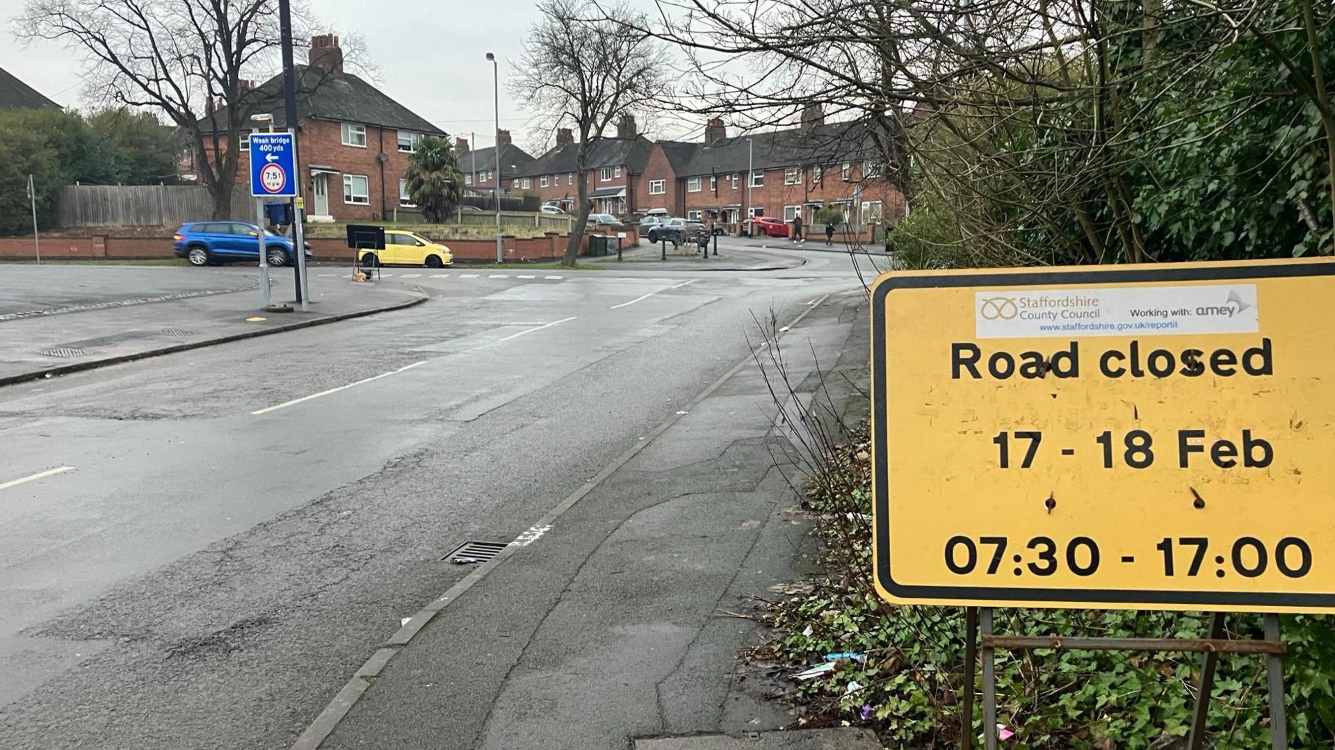 A yellow road sign with details of a road closure, is positioned at the side of a road with a junction visible in the distance.