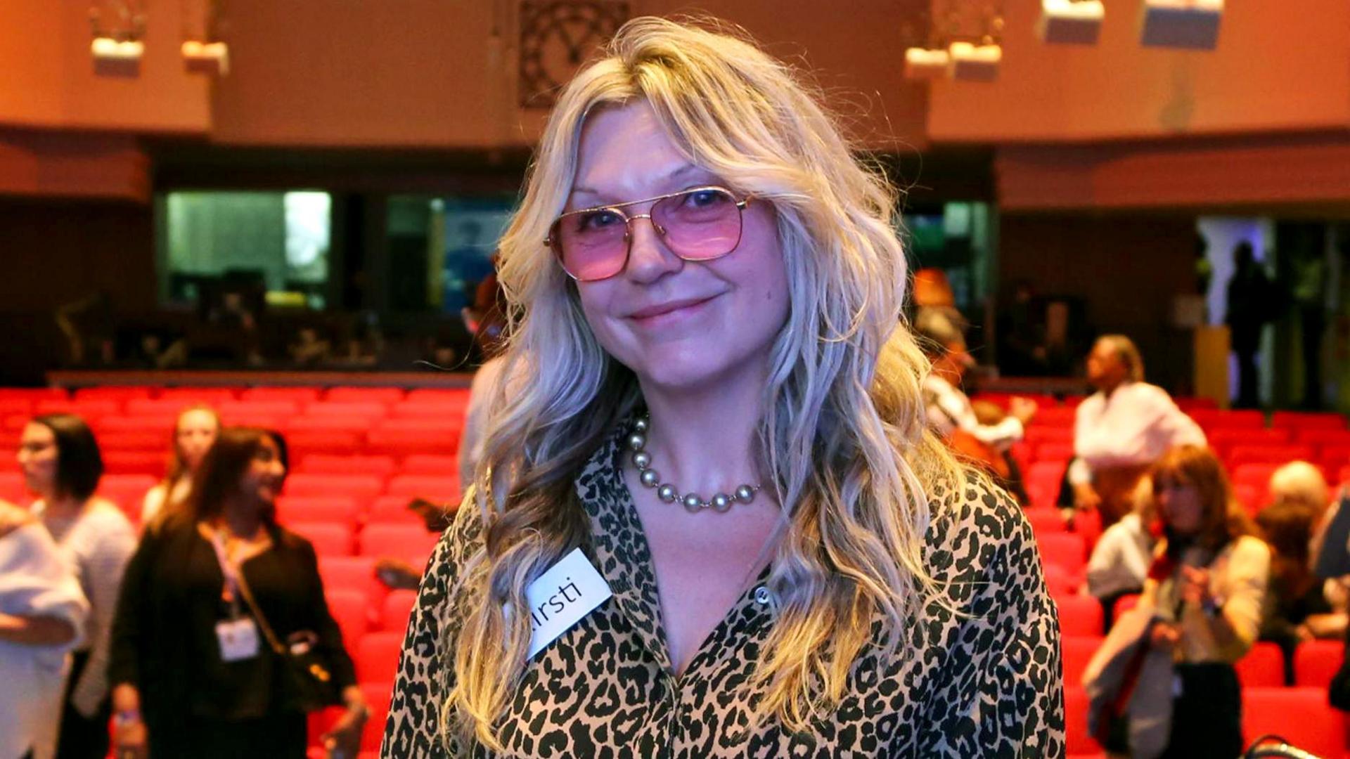 Kirsti wears pink-tinted glasses and a leopard print shirt. She is smiling into the camera while in wht looks like an auditorium with people and red seats in the background