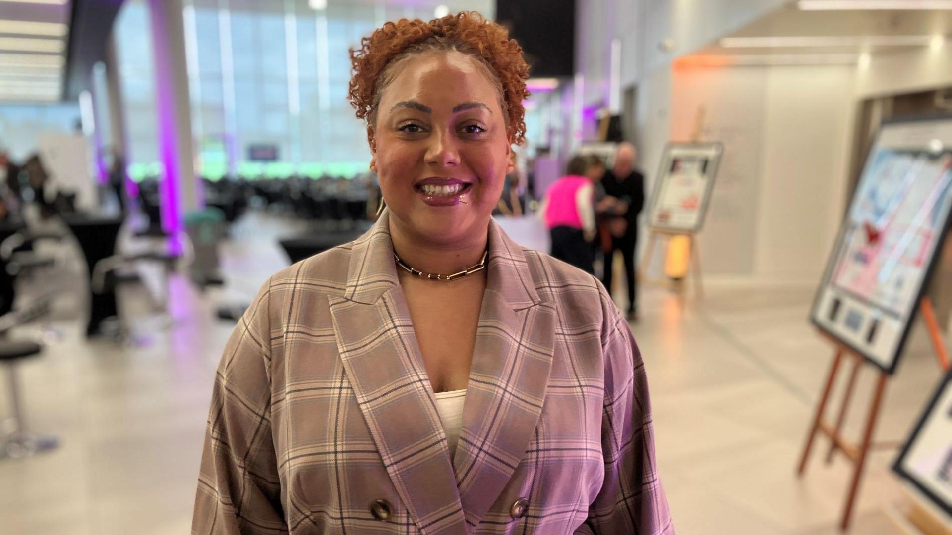 Campbwell stands indoors in a bright, modern space with large windows behind her. She is wearing a plaid blazer cinched at the waist with a white belt and has curly hair. In the background, several other people are visible, some of whom are observing framed displays mounted on easels.