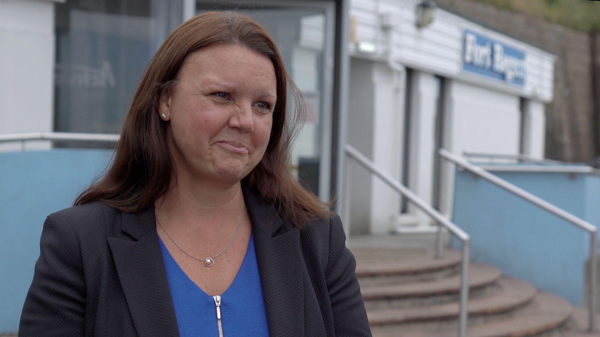 Emma Baker looking to the right and standing in front of a building. She has brown hair and is wearing a blue top with a black blazer over the top of it. In front of the building are steps leading to the entrance.