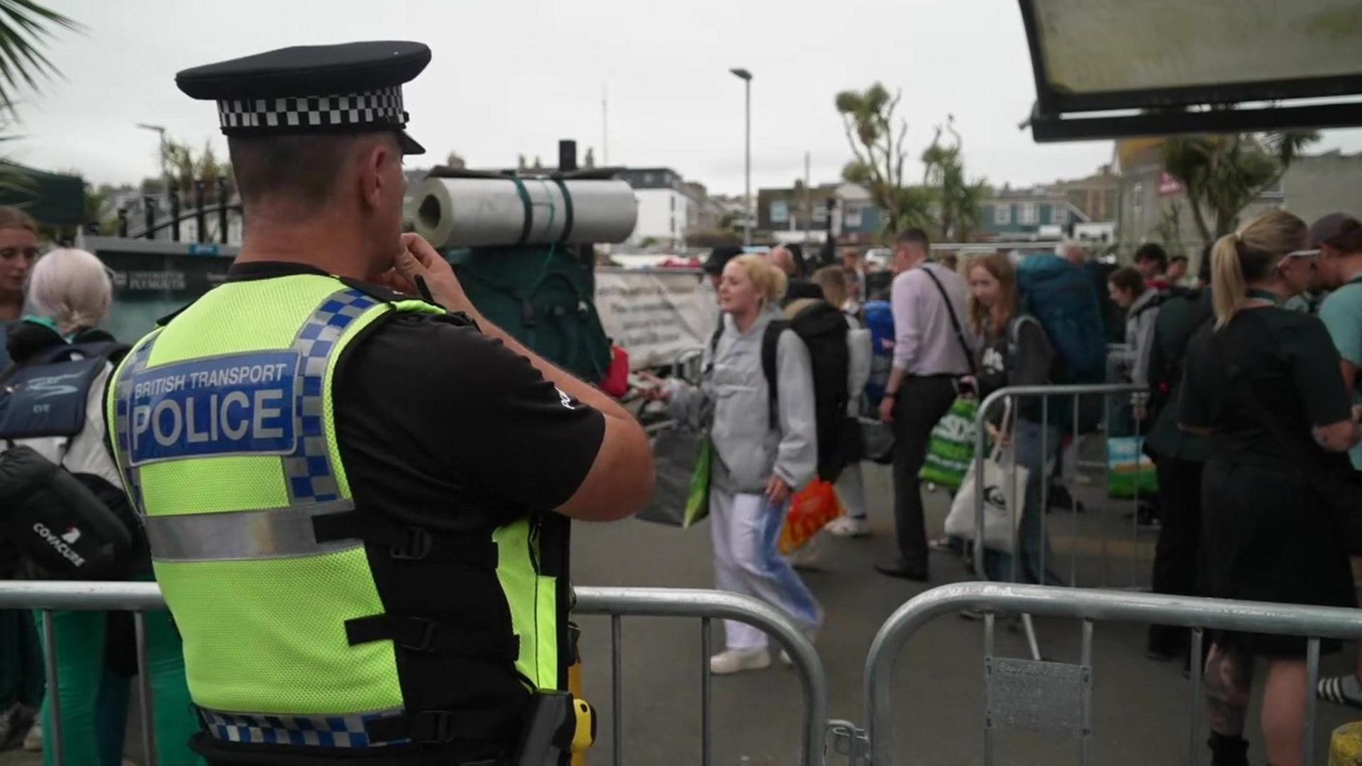 Back of male police officer wearing a yellow high-viz vest looking out on to approximately 30 people holding bags and items for packing, grey weather