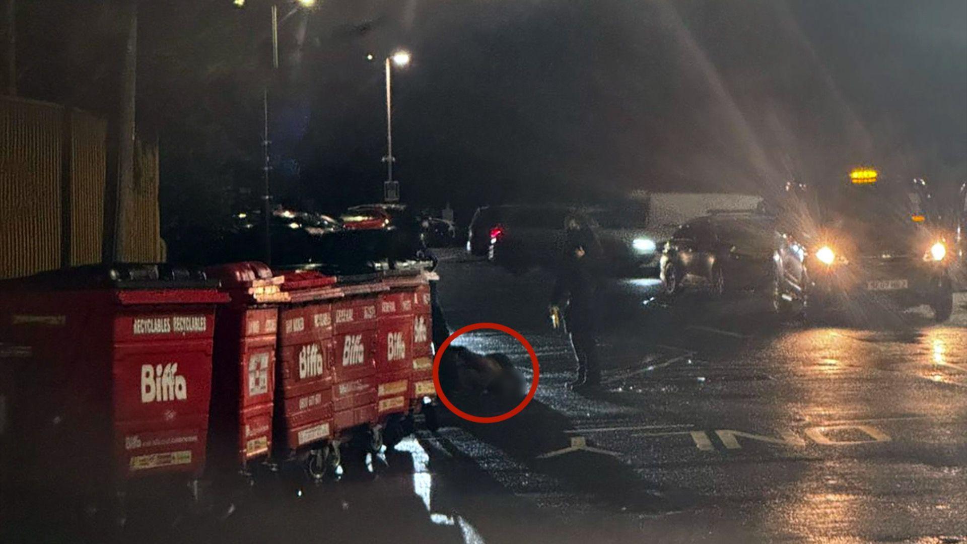 Një imazh i errët i një parkingu makinash. Një rreth i kuq përshkruan imazhin e turbullt të një burri në tokë. Një oficer qëndron në të djathtë.