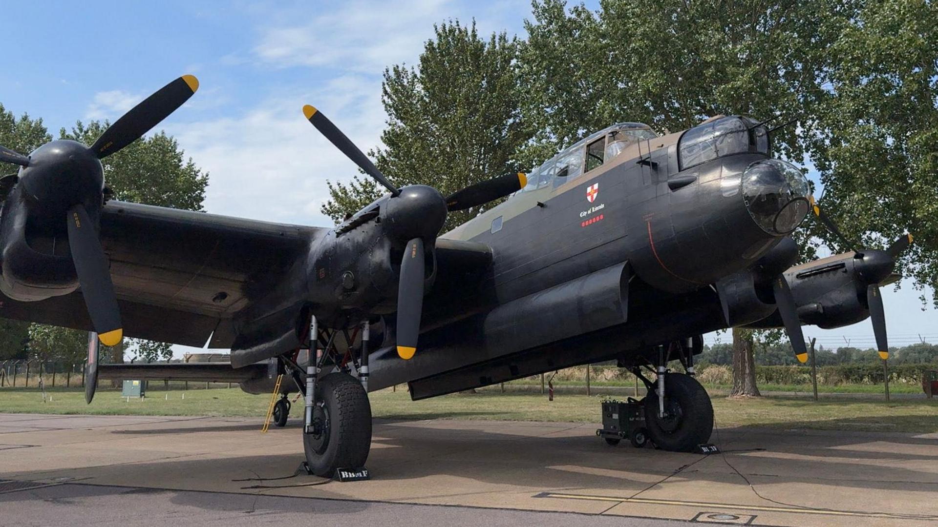 The photo shows a black Lancaster Bomber aircraft. It's stationary, and is sat on a runway. There are trees in the background.