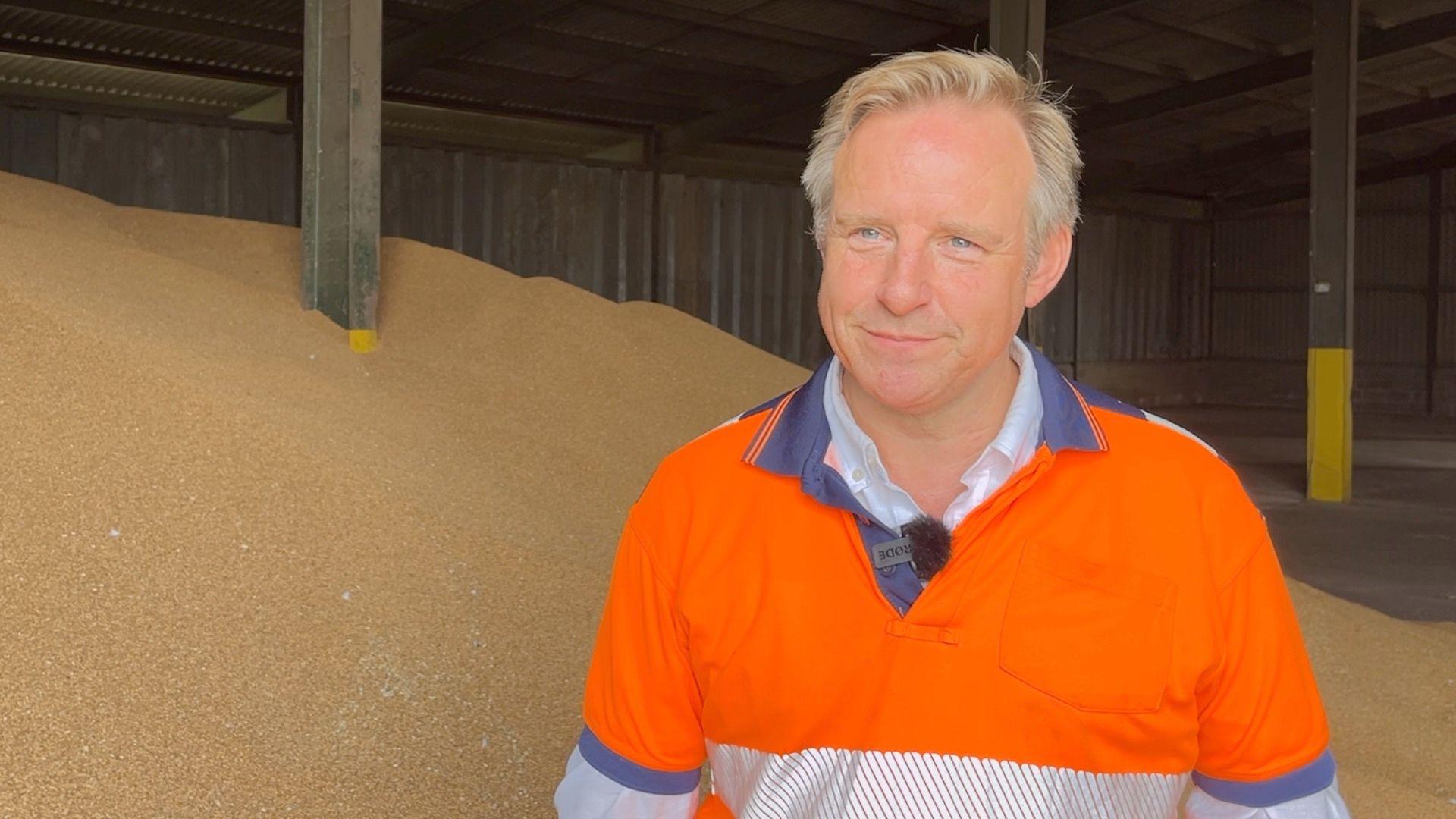 Kit Papworth is standing in front a pile of grain. He is wearing orange high-visibility top. He has blonde greying hair.