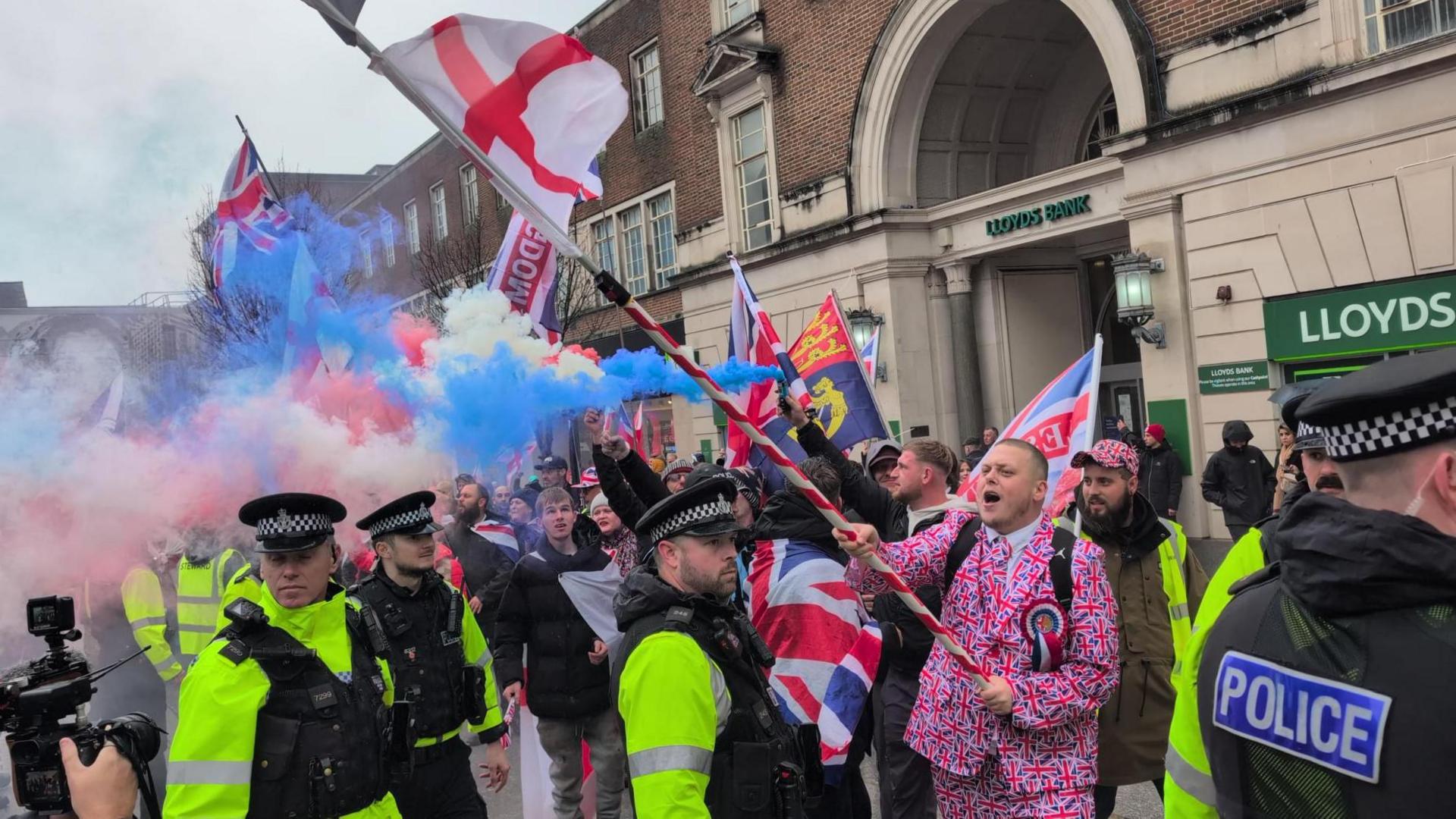 Protesters waving England and Union flags stand in front of police. Red, white and blue smoke flares have been set off. 