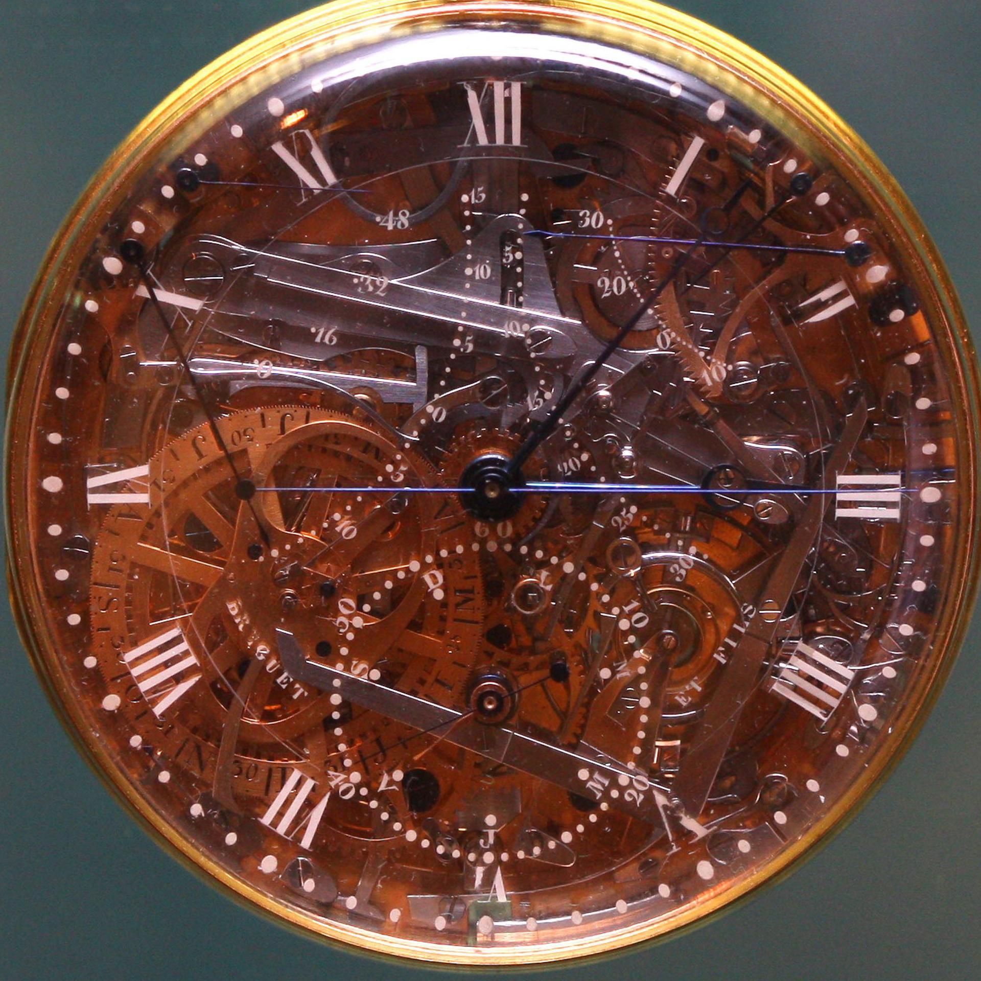 The gold casing of the watch with white roman numerals and a transparent crystal dial, showing hundreds of tiny platinum and gold parts underneath