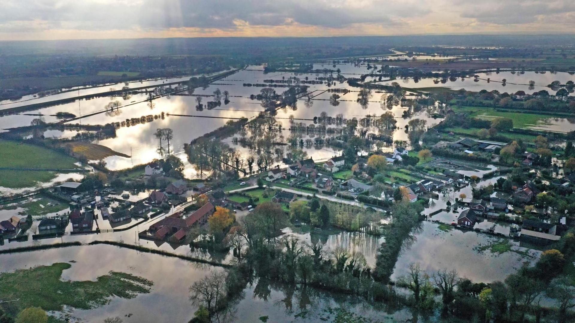 A bird's eye view of a houses and fields surrounded by flood water stretching out to the horizon
