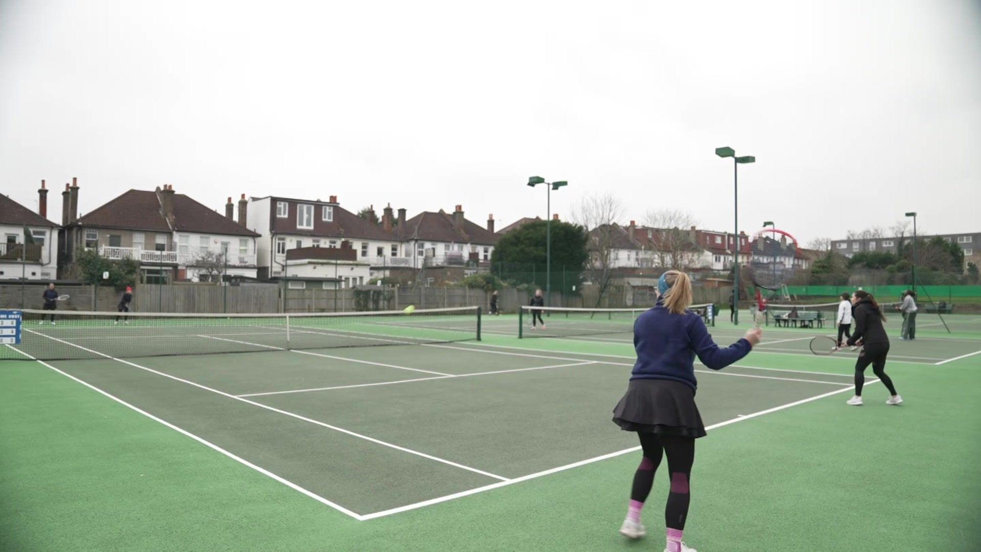 Two women with tennis rackets about to hit a green ball at the back of a green court with two women the other side of the net