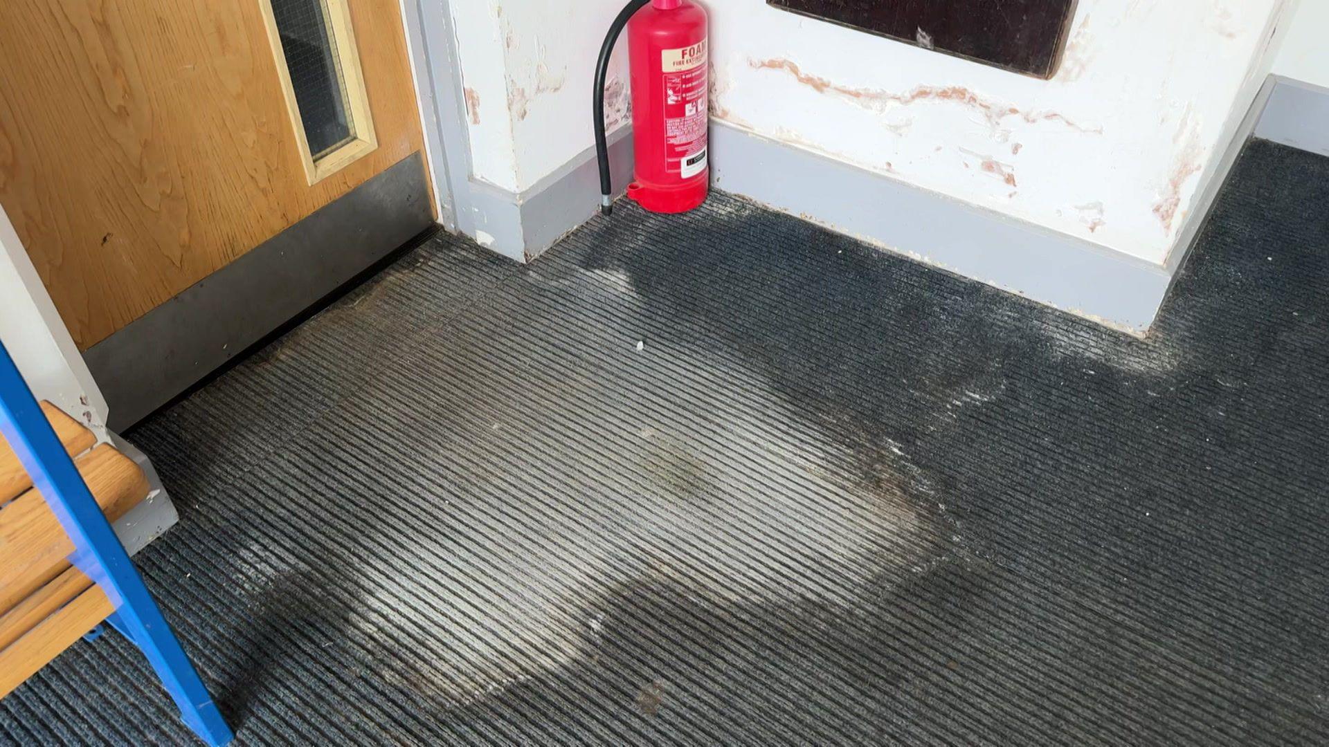 A close-up of a blue school corridor carpet with a large water damage stain emerging from the bottom of a door. The wall nearby also shows signs of water damage, and a fire extinguisher is visible in the corner.