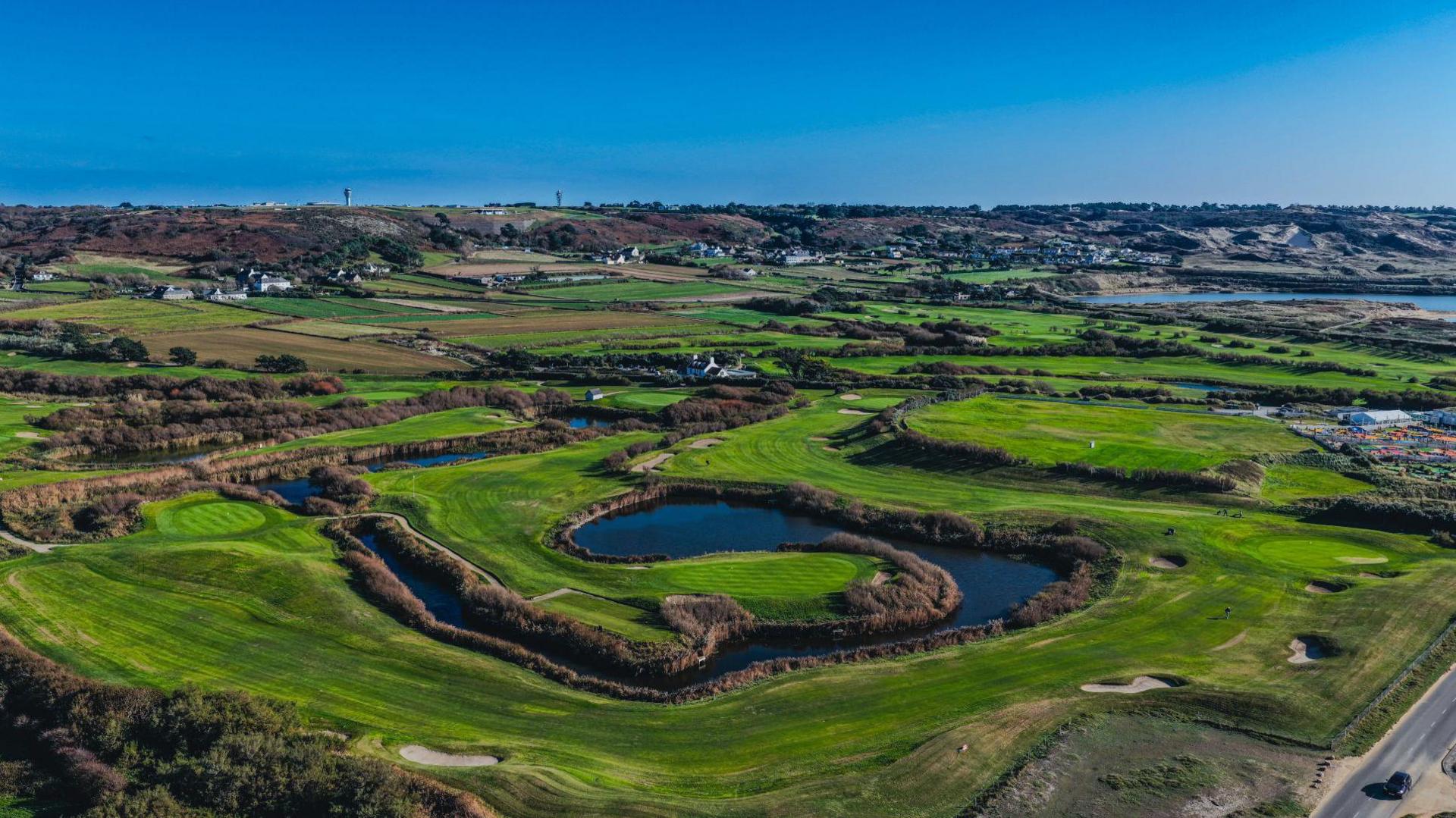 A green golf course is besides the sea and has a large pool of water in the centre.